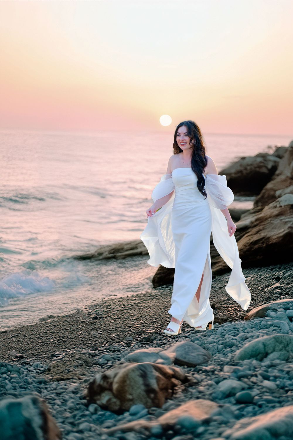 Woman in flowing white dress walks along rocky beach shore at sunset with golden sun and pink sky over ocean.
