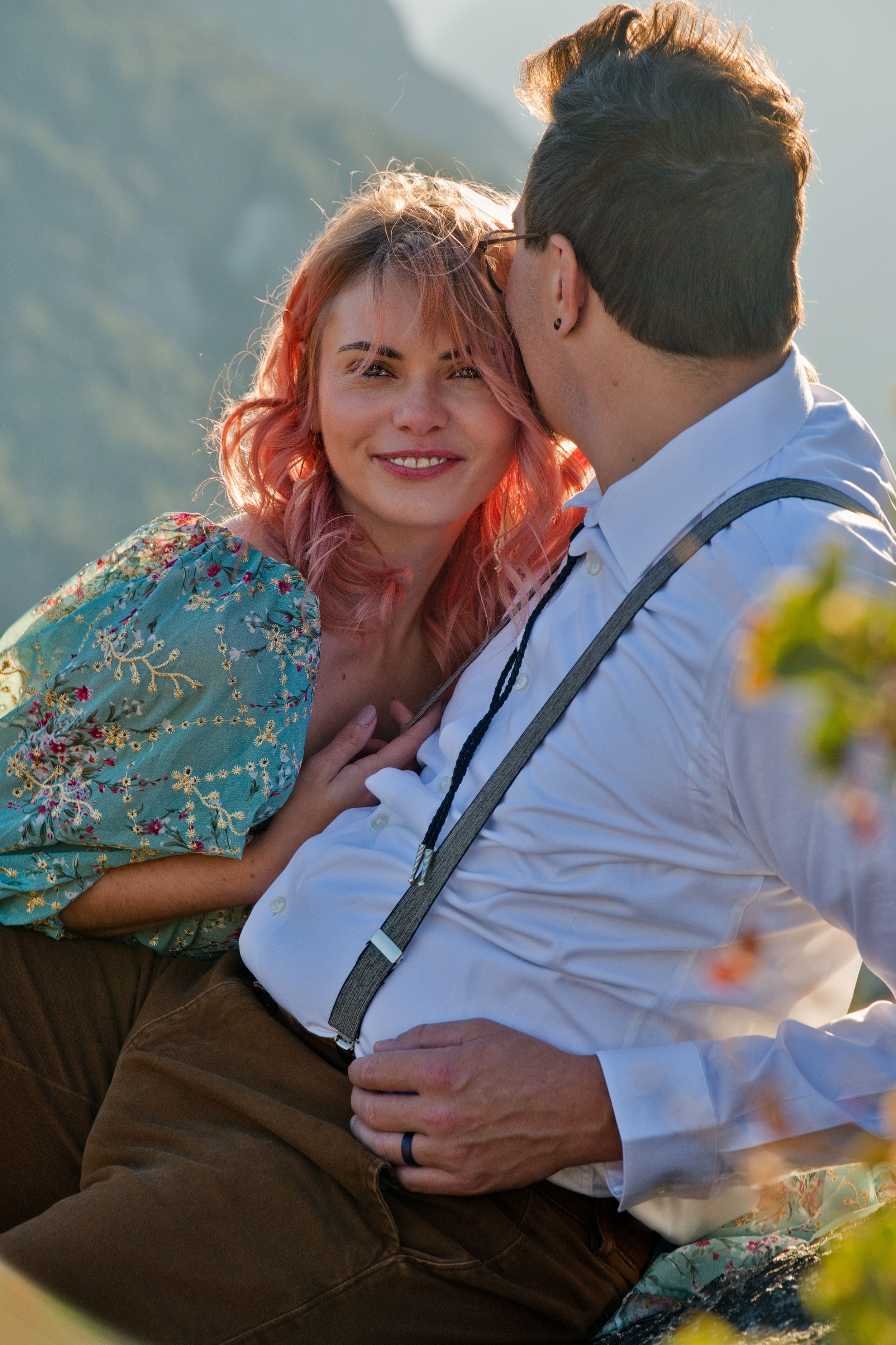 A romantic couple sits together outdoors, with a woman in a floral dress and a man wearing suspenders against a mountain backdrop.