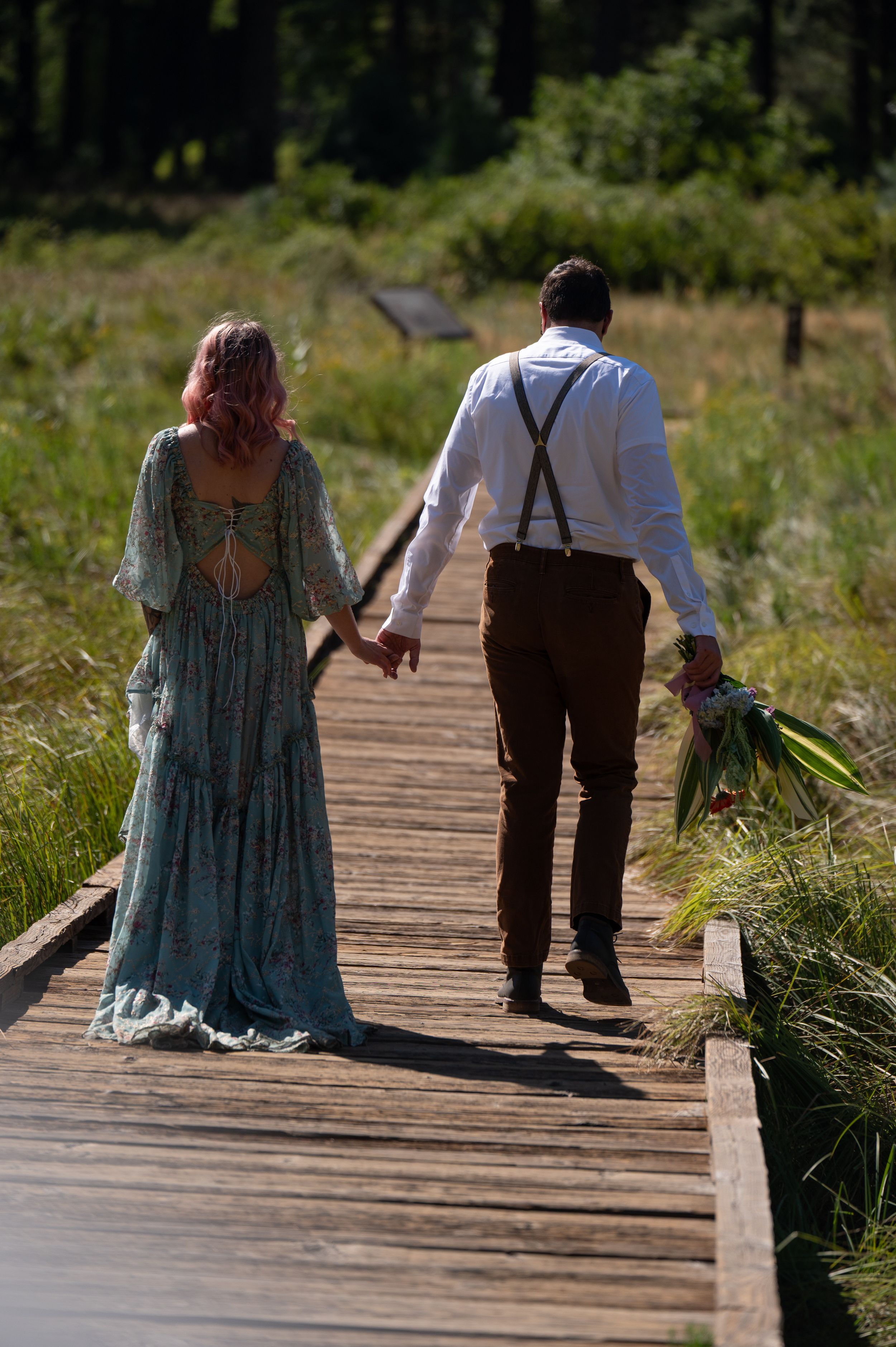 A couple walks hand in hand down a wooden boardwalk path, with the person in suspenders holding a bouquet.