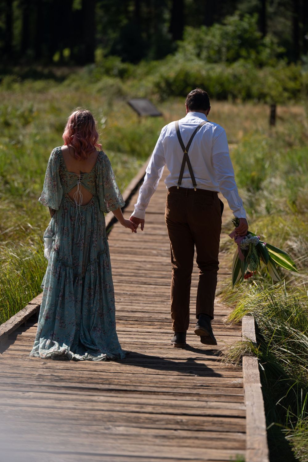 A couple walks hand in hand down a wooden boardwalk path, with the person in suspenders holding a bouquet.
