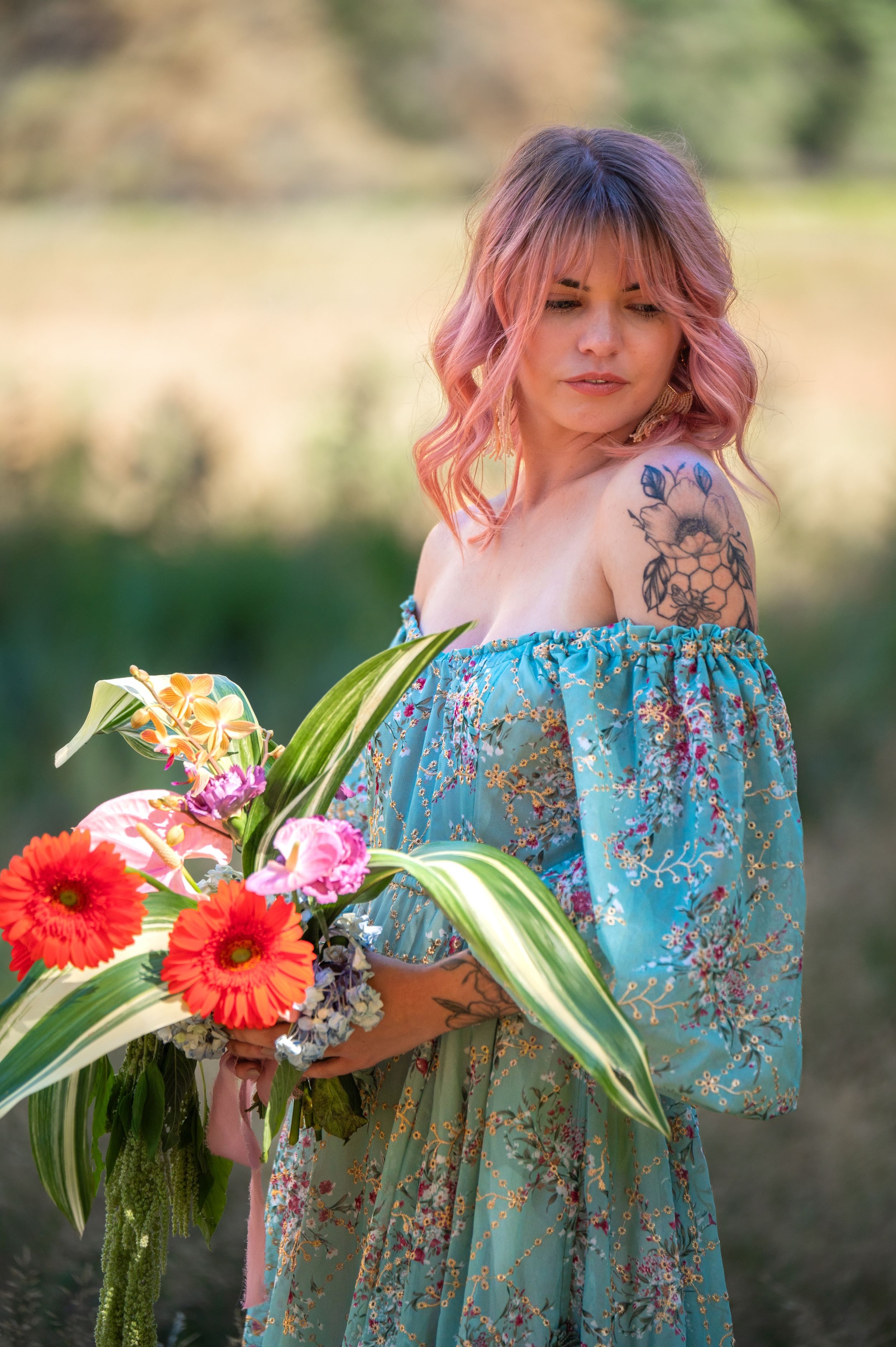 Someone in a blue floral off-shoulder dress holding colorful flowers with gerbera daisies stands in a dreamy outdoor setting.