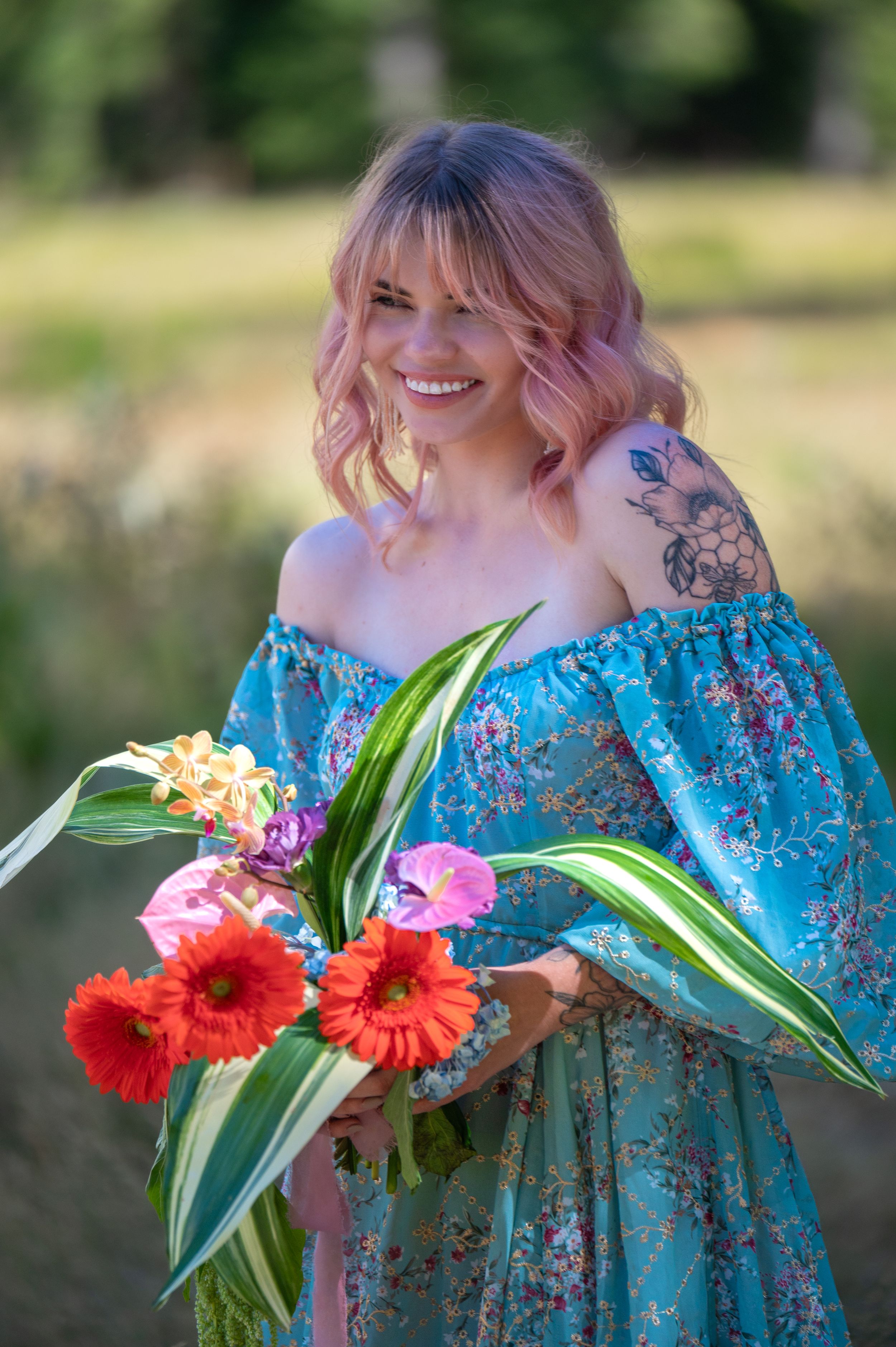 A bright portrait featuring pastel pink hair, a blue floral dress, and colorful red gerbera daisies with tropical foliage.