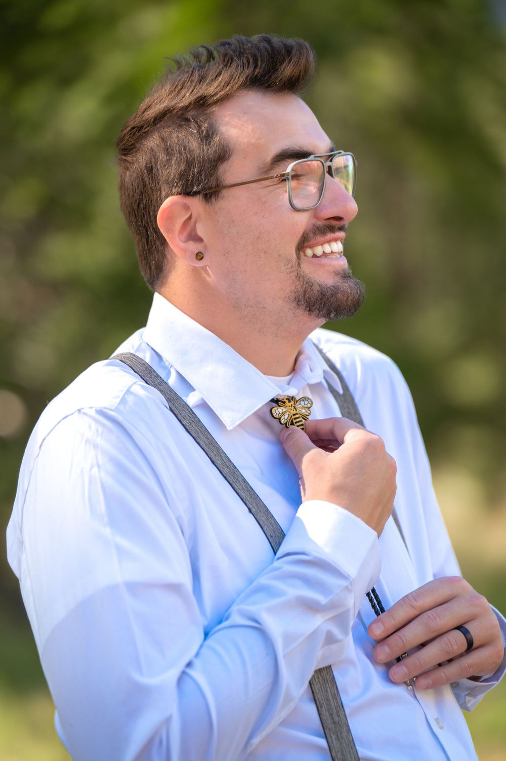 A groom in suspenders and a white dress shirt smiles while adjusting his butterfly-shaped boutonniere.