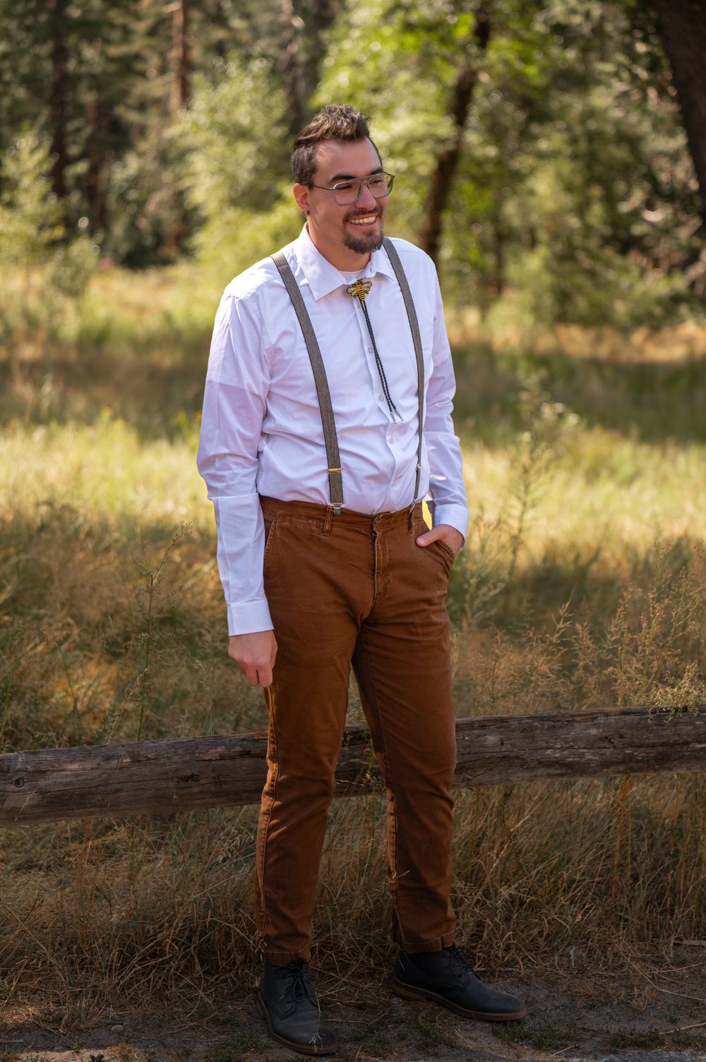 A person wearing suspenders and brown pants smiles while standing in a sun-dappled meadow.