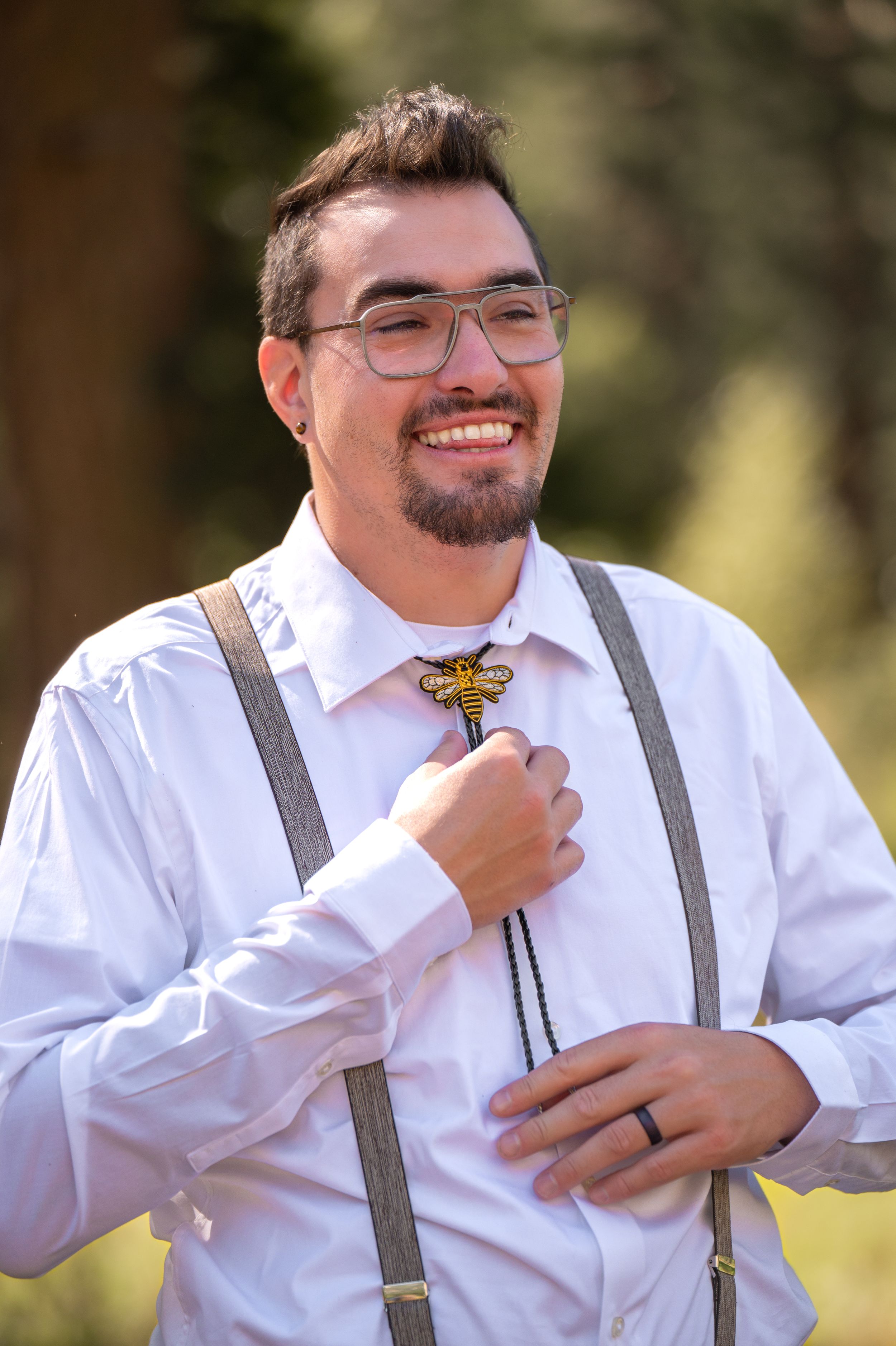 A happy groom wearing suspenders and glasses touches his unique butterfly-shaped lapel pin.
