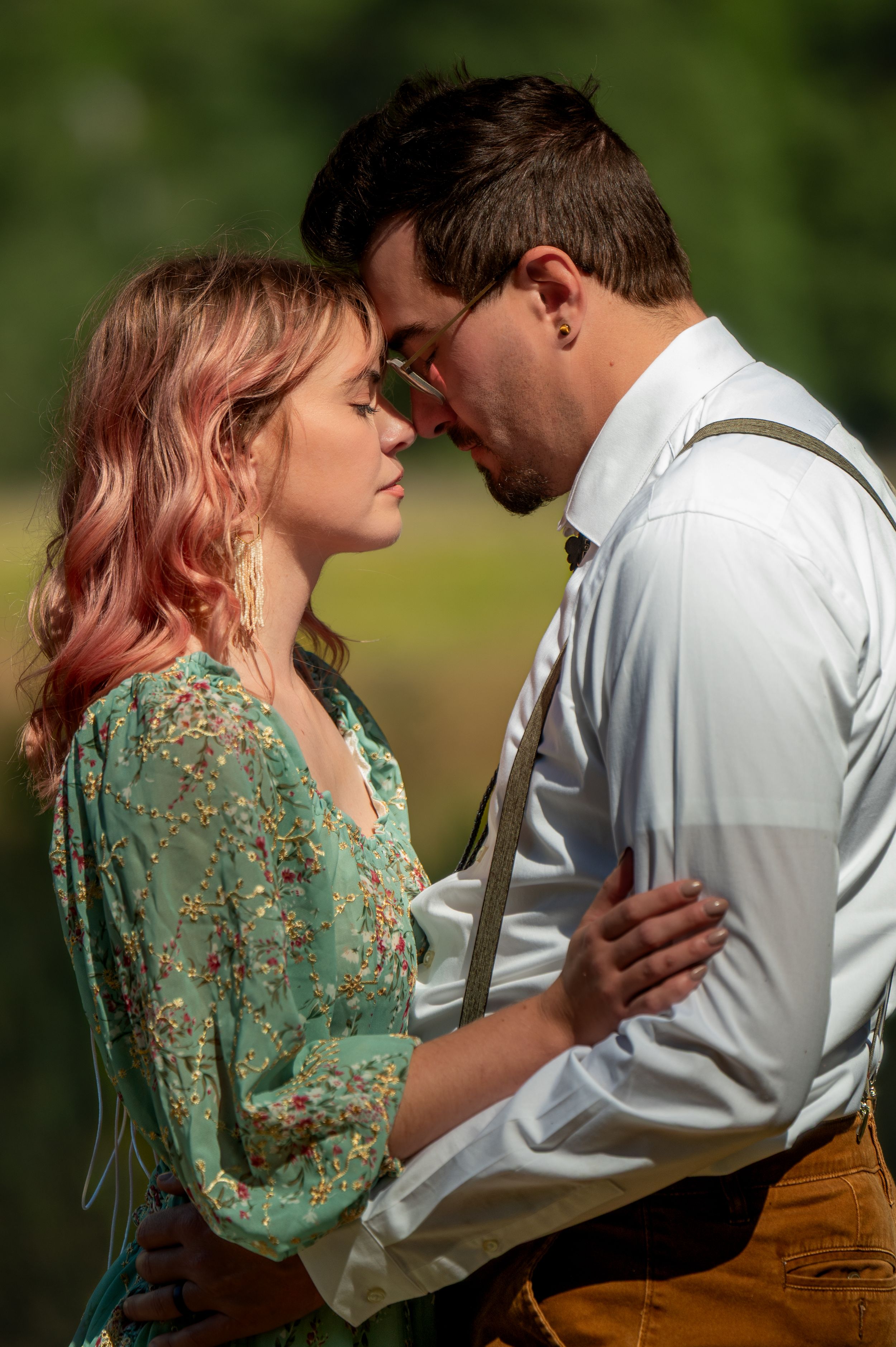 A romantic portrait in nature showing a couple in an intimate embrace, with her in a green floral dress and him in suspenders.
