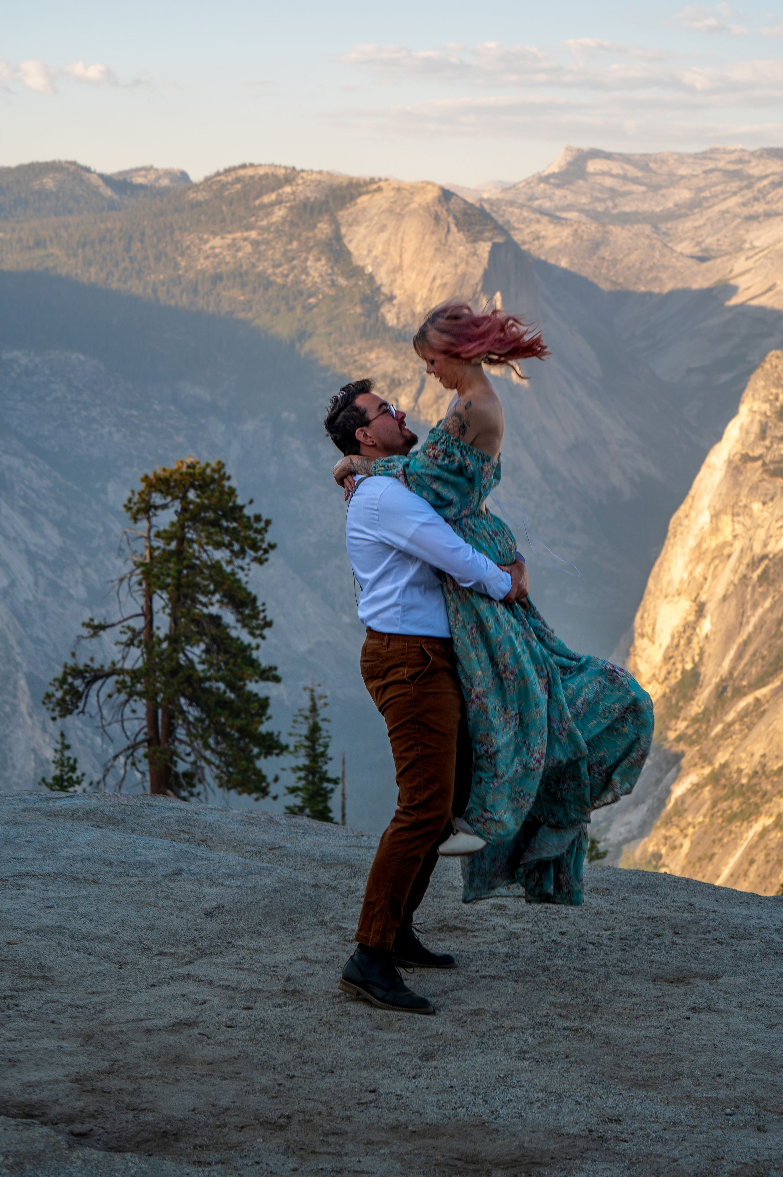 A romantic couple embraces on a cliff edge overlooking majestic Yosemite Valley mountains at sunset.