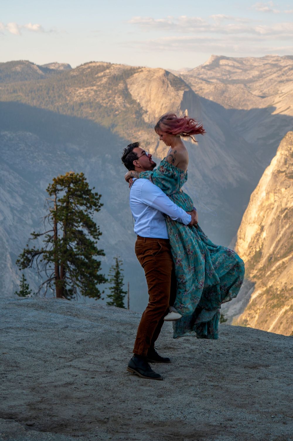 A romantic couple embraces on a cliff edge overlooking majestic Yosemite Valley mountains at sunset.