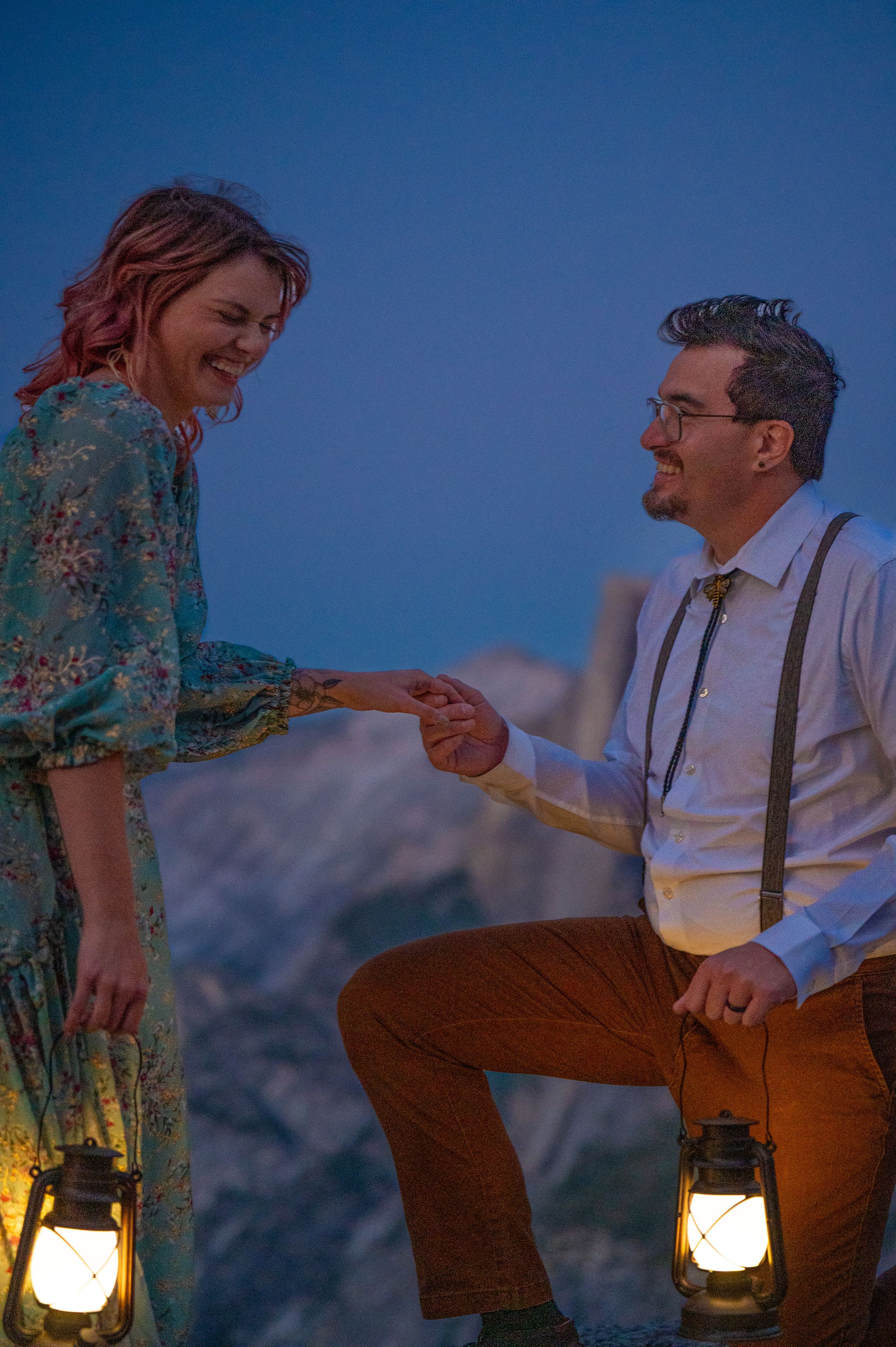 A couple shares a romantic moment at dusk with lanterns glowing against a mountain backdrop.