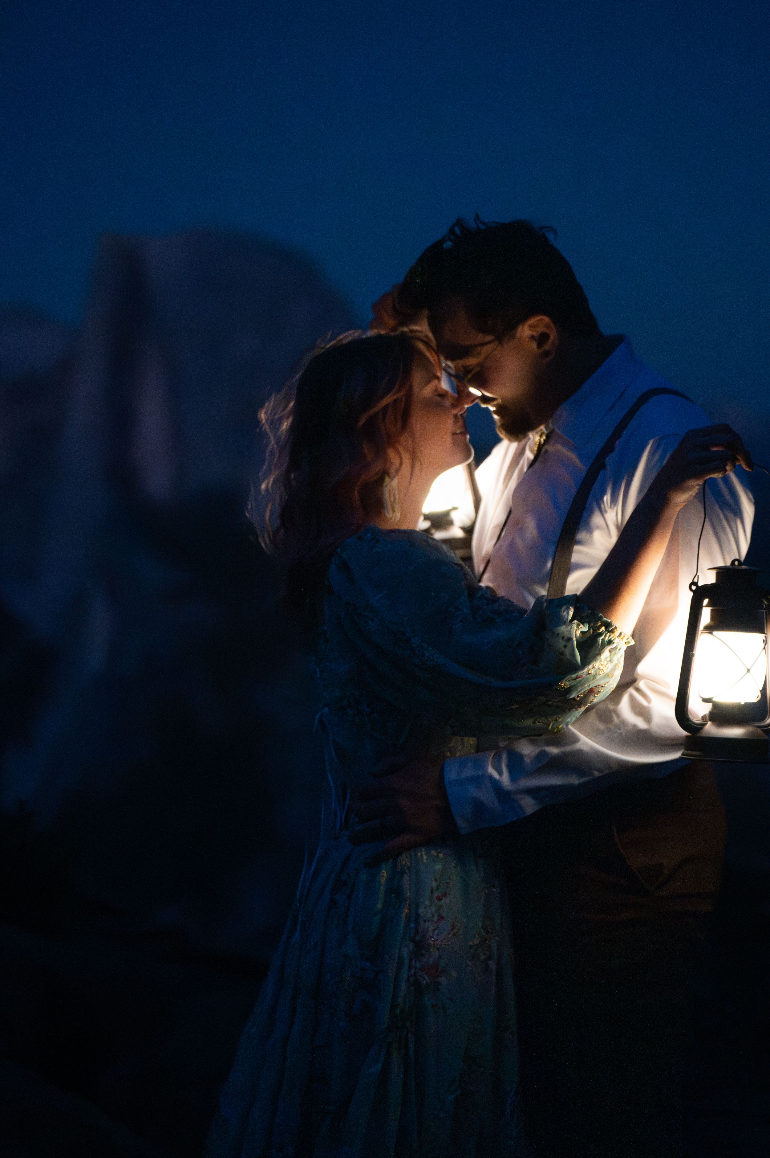A couple shares an intimate moment under lantern light with dramatic mountain silhouettes in the background.