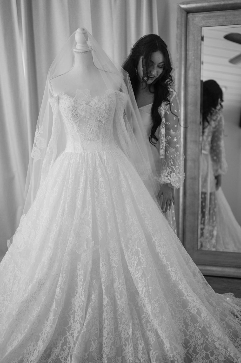 A flowing white lace wedding dress with long sleeves and veil displayed on a mannequin beside a mirror.