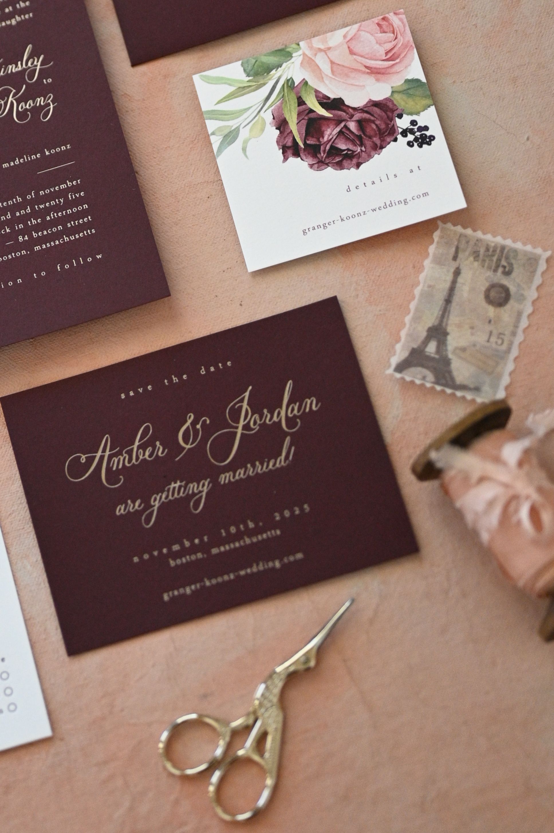 Burgundy wedding invitation suite with floral design, vintage stamps and gold scissors on a blush pink background.