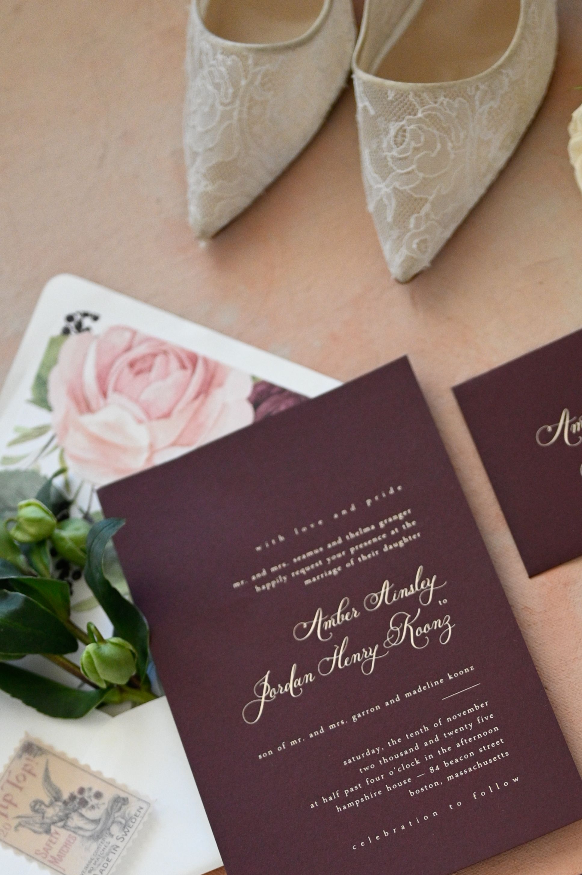 Elegant lace wedding shoes and burgundy invitation suite with pink florals displayed on light peach background.