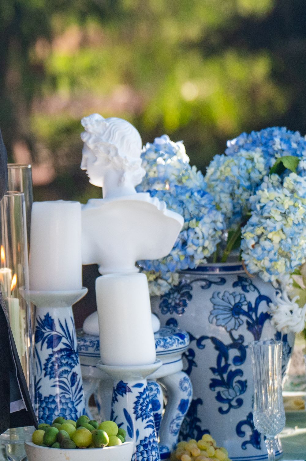 Elegant table setting with blue hydrangeas in chinoiserie vases, white candles, and a classical bust statue in soft lighting.