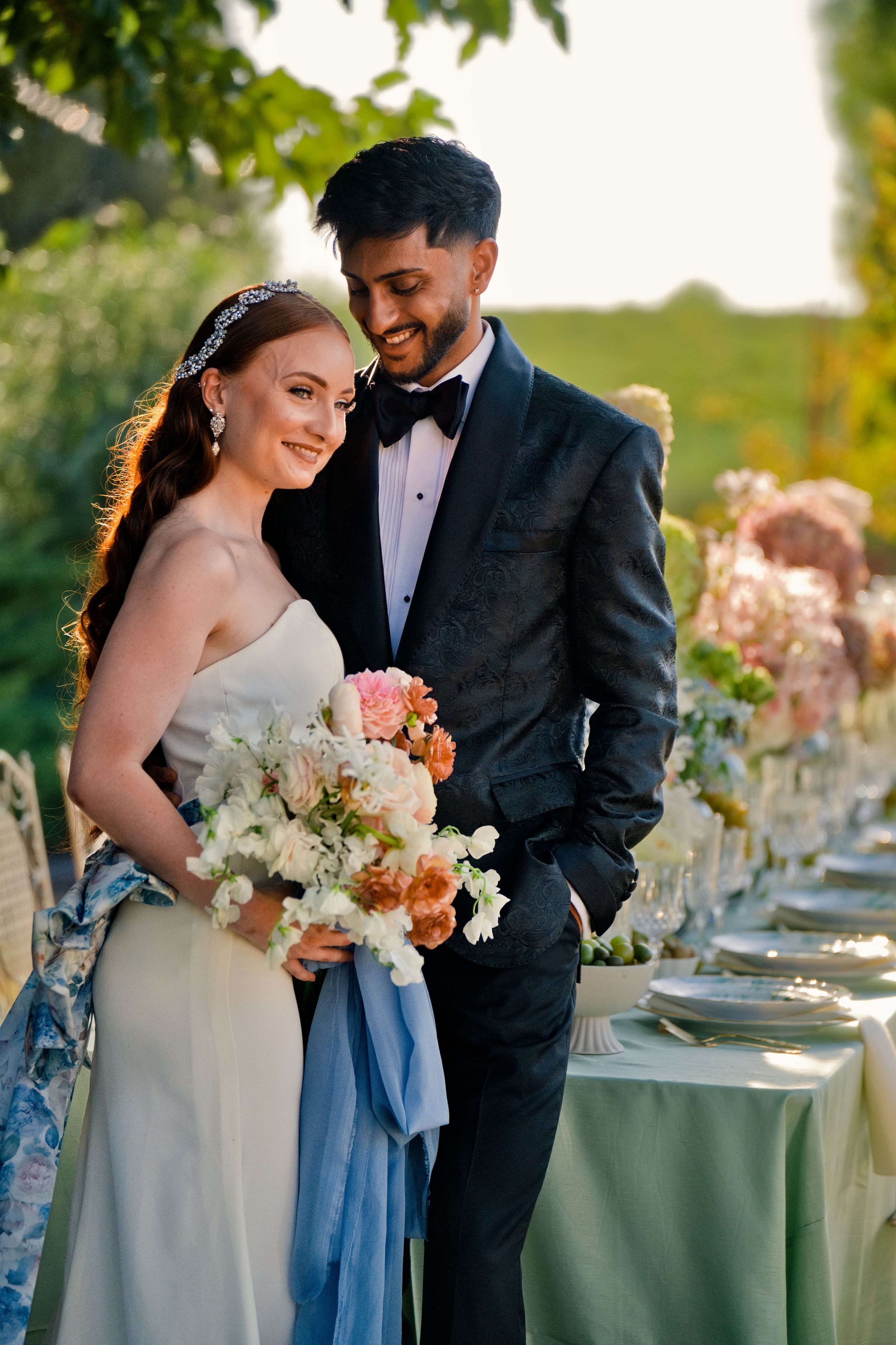 A happy couple shares a moment beside their elegant outdoor wedding reception table decorated with fresh flowers and linens.