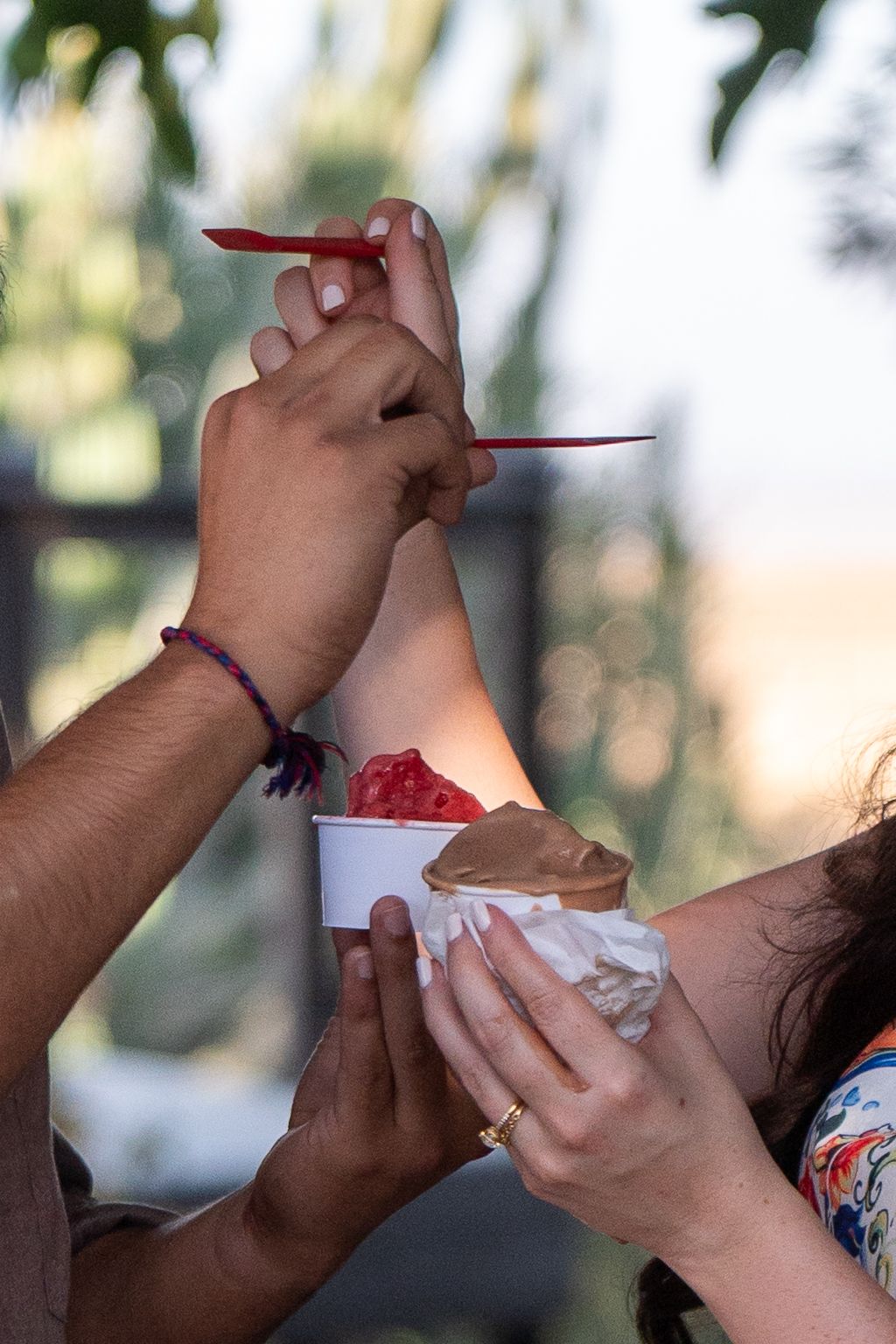 Hands holding shaved ice dessert with red syrup being served at an outdoor gathering in natural lighting.