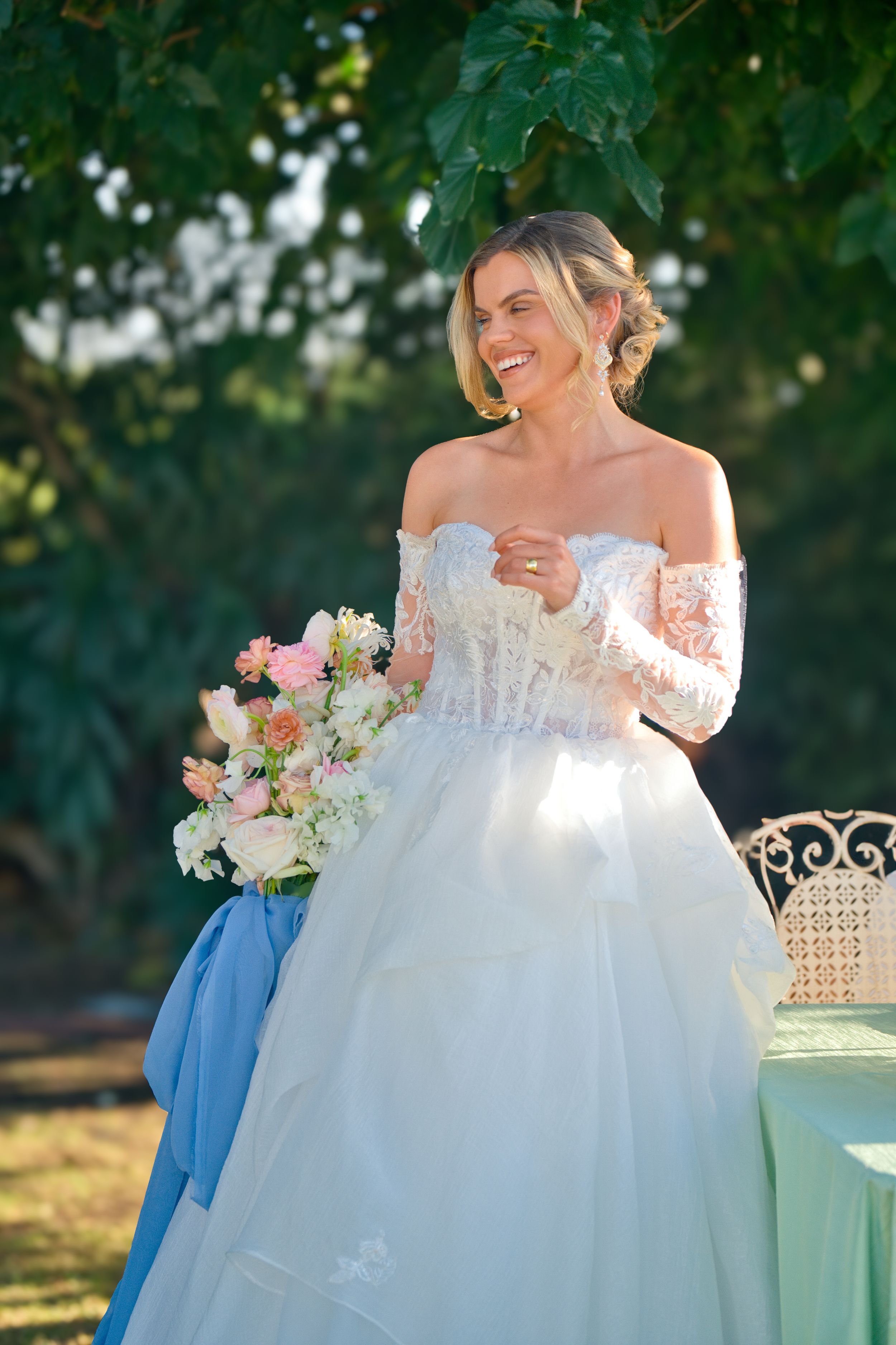 A bride in an off-shoulder lace gown holds a pink and white bouquet while standing outdoors under green foliage.