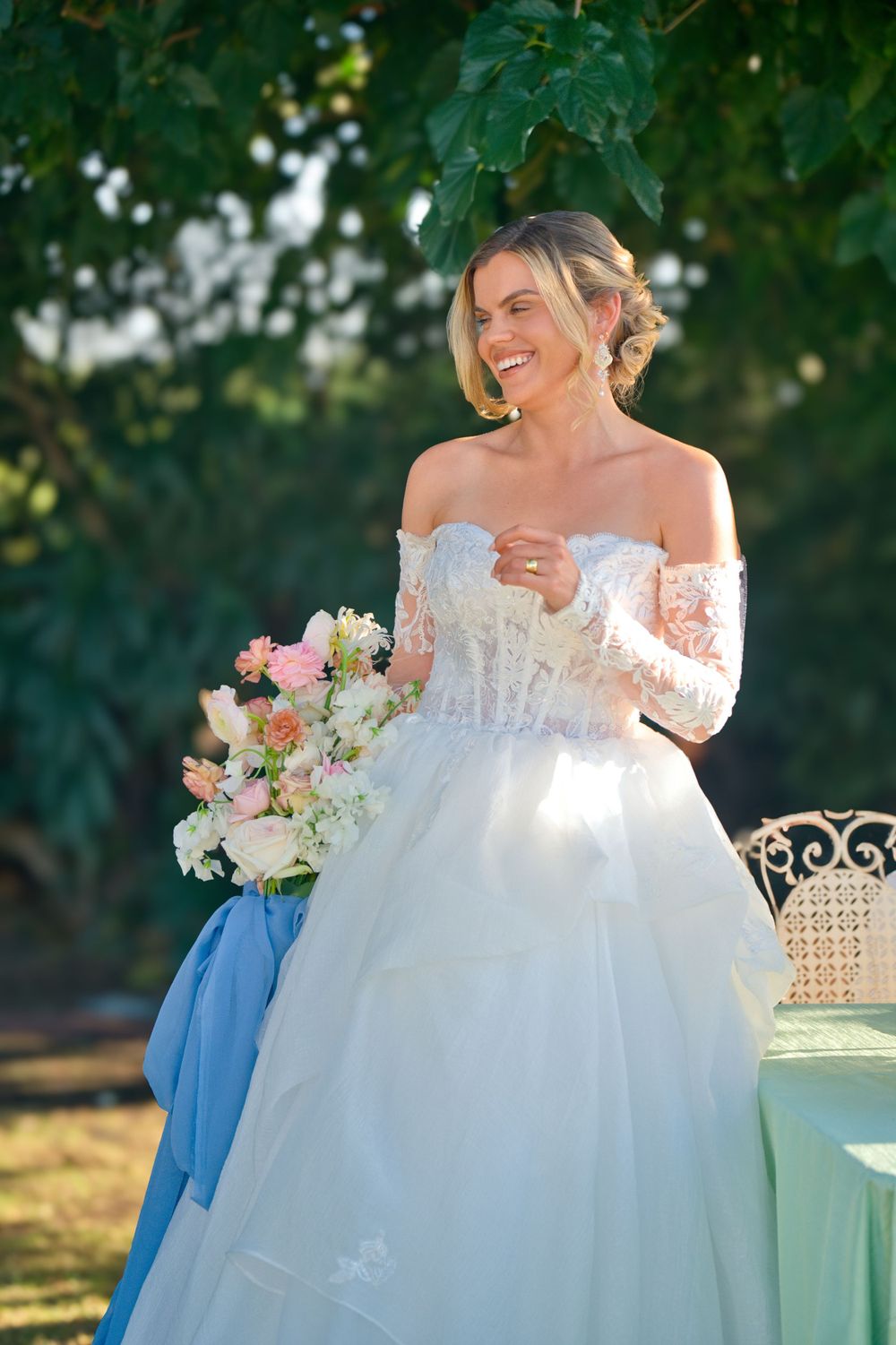 A bride in an off-shoulder lace gown holds a pink and white bouquet while standing outdoors under green foliage.