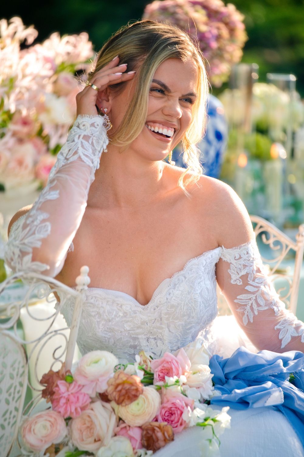A radiant bride in a lace off-shoulder wedding gown laughs joyfully while holding a bouquet of pink and white roses.