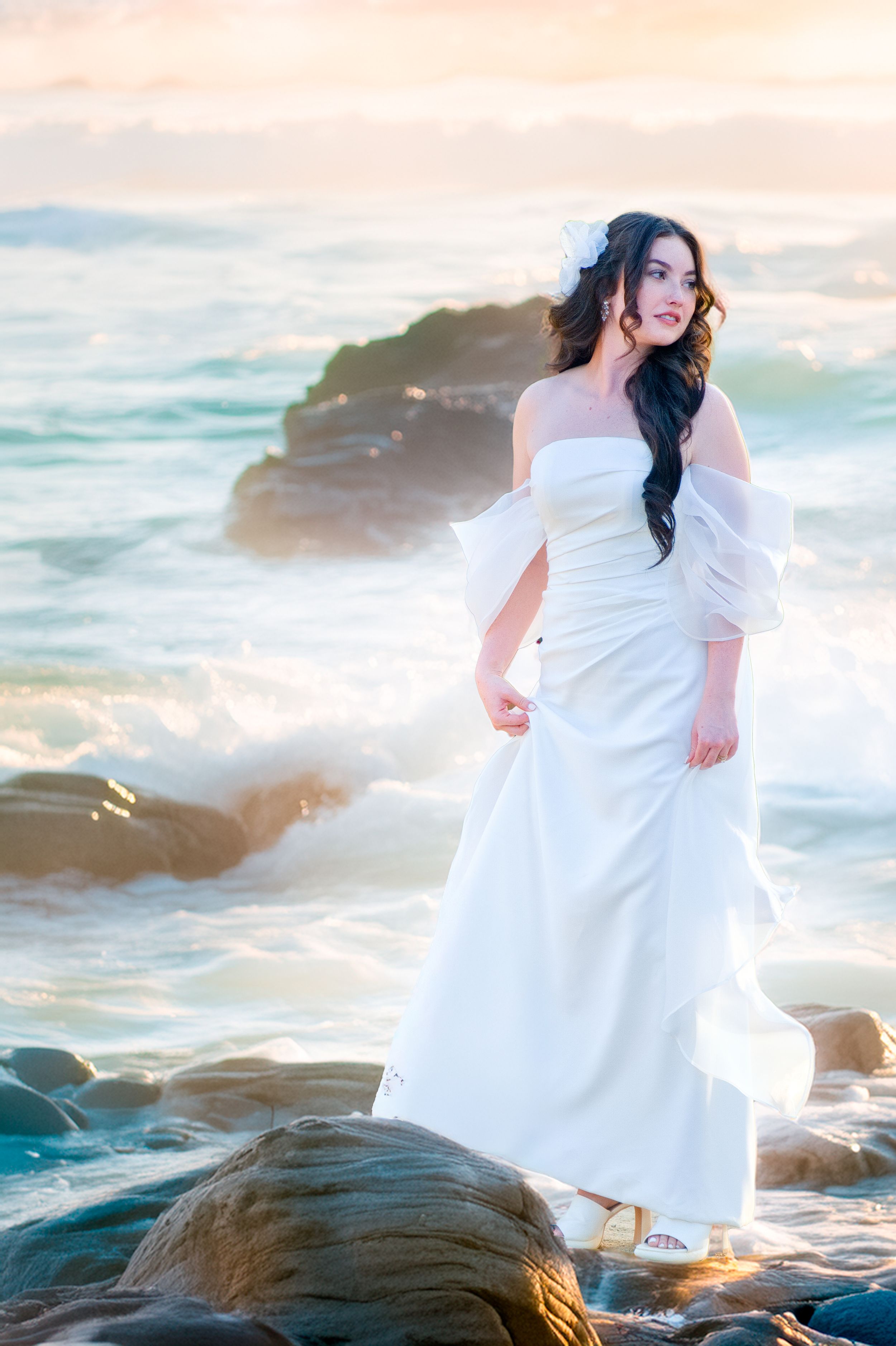 A person in a flowing white off-shoulder dress poses on coastal rocks at sunset with waves crashing in the background.