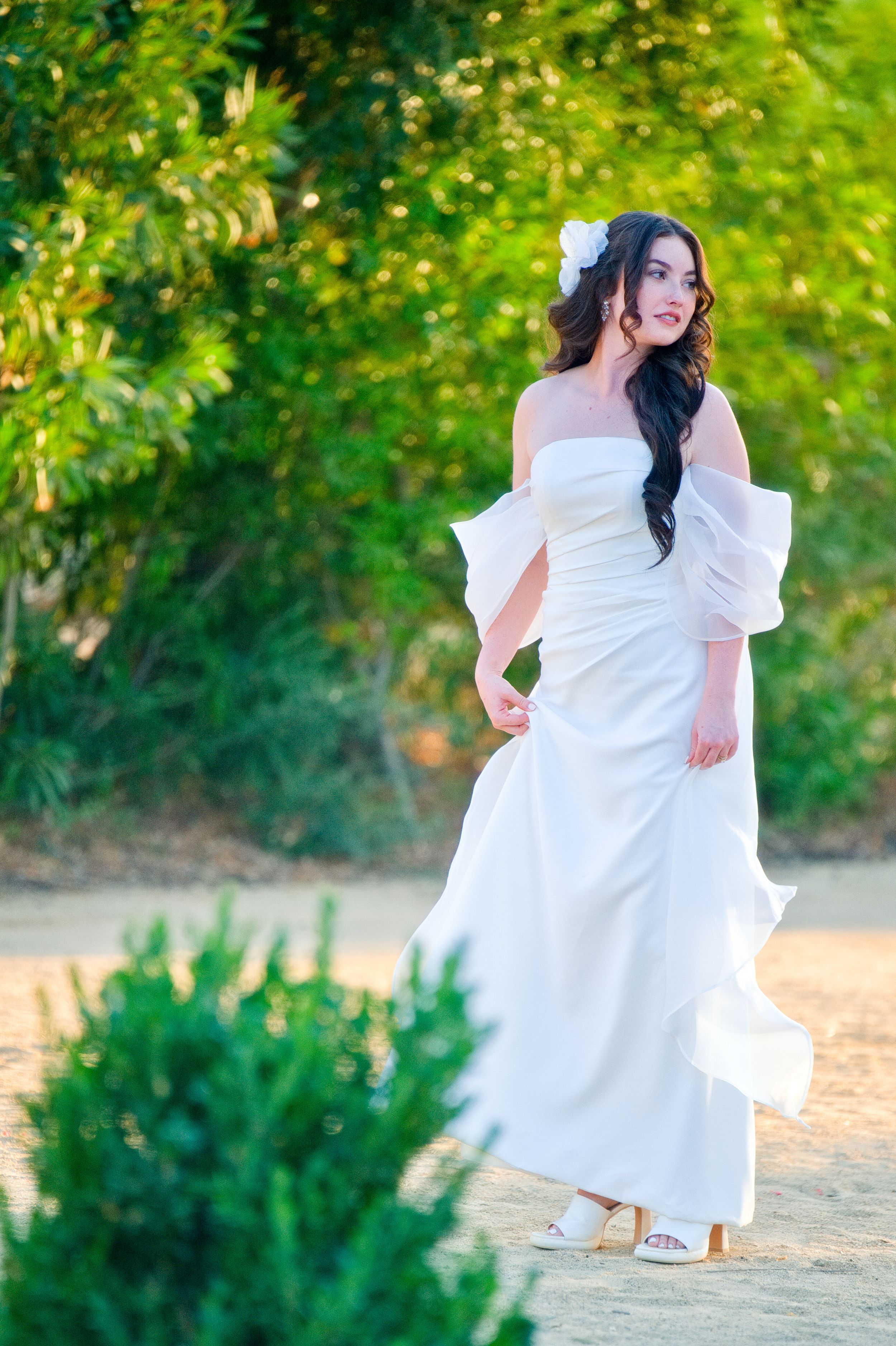 Elegant woman in flowing white off-shoulder dress poses gracefully on a nature path with sunlit foliage in the background.