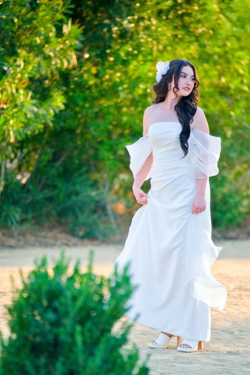 Elegant woman in flowing white off-shoulder dress poses gracefully on a nature path with sunlit foliage in the background.