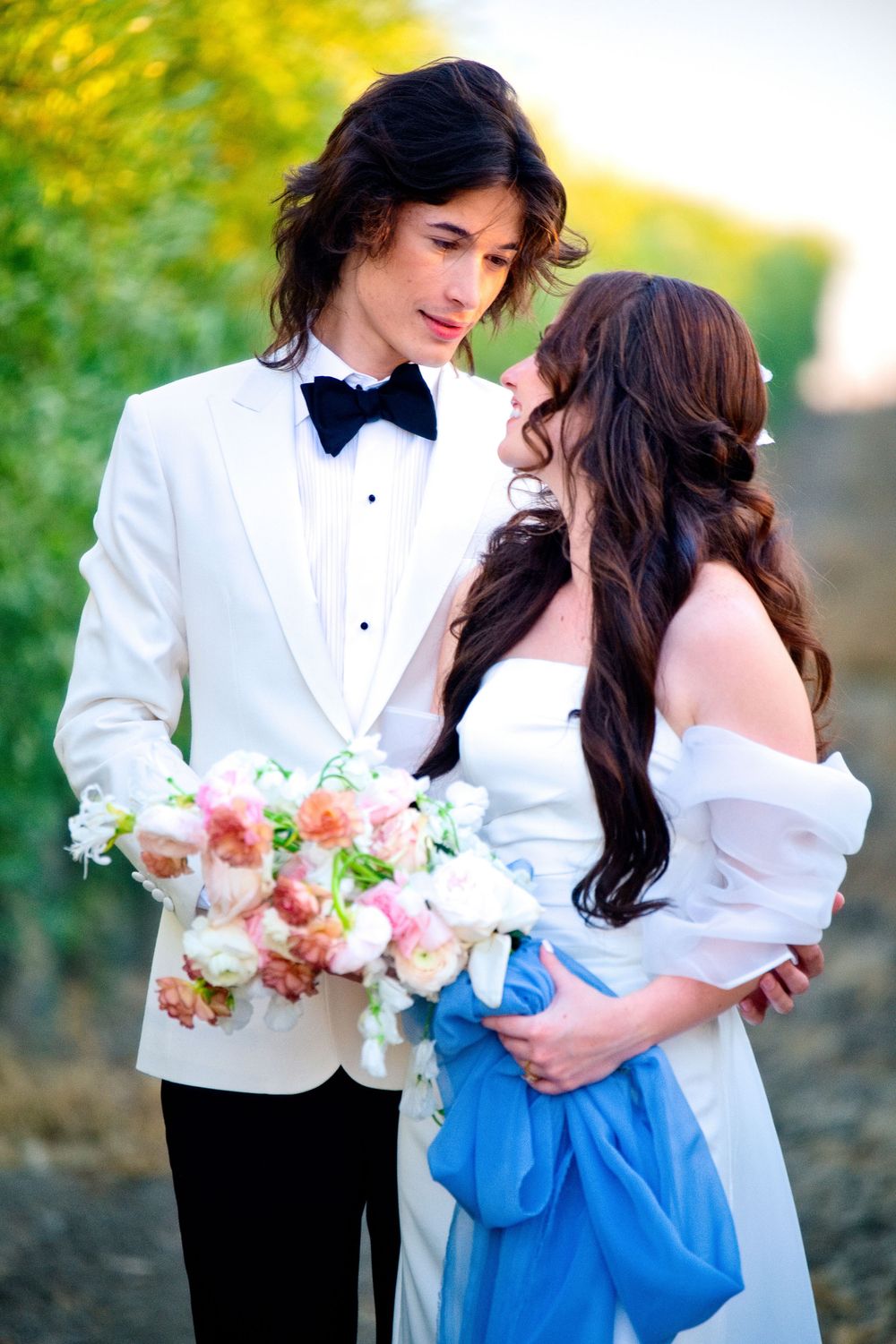 A bride and groom share a romantic moment outdoors with peach florals and white formal attire.
