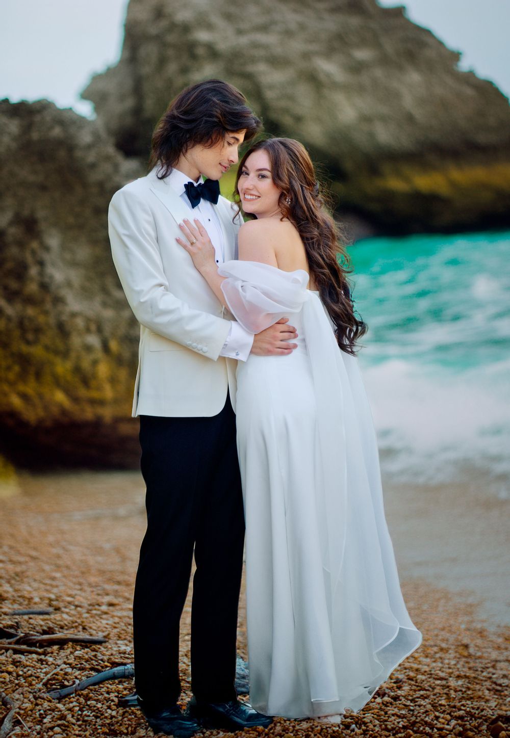Romantic couple embraces on a beach with turquoise water and rocky cliffs, dressed in formal white wedding attire.
