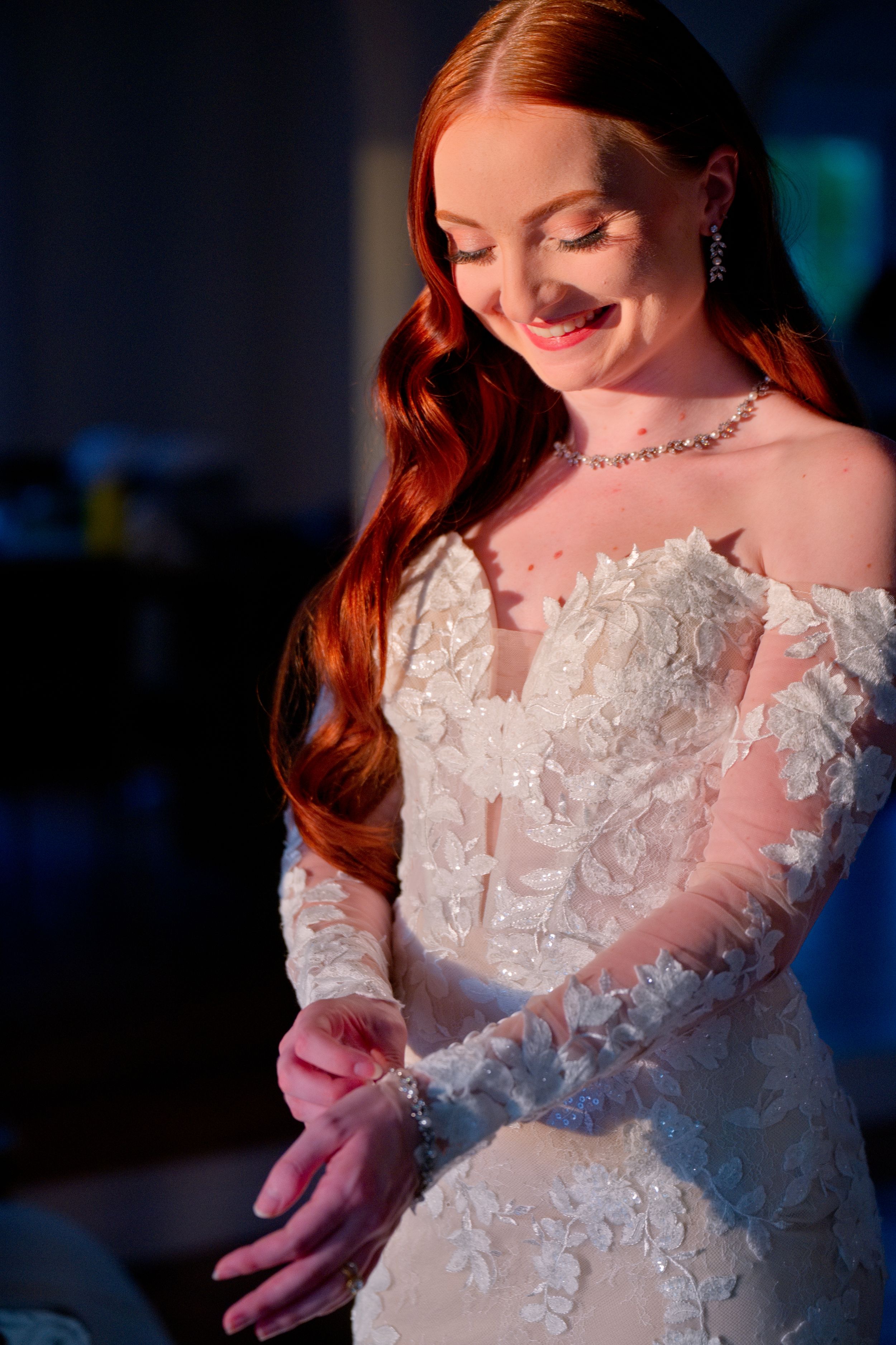 A bride in an ornate lace wedding gown with off-shoulder sleeves and long red hair smiles while looking down.