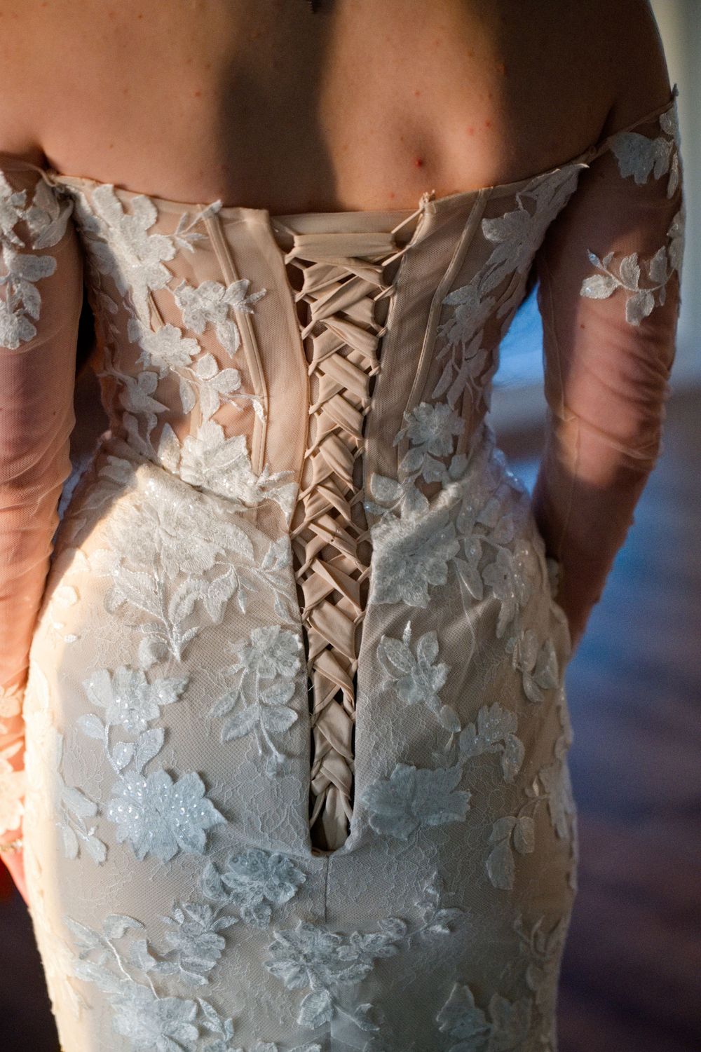 Close-up detail of a white lace wedding dress showing intricate floral embroidery and corset-style lacing on the back.