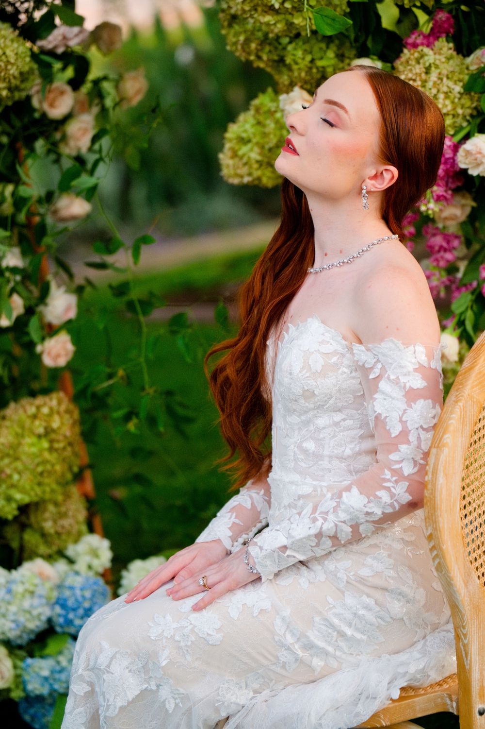 A romantic garden portrait of someone in a white lace wedding dress with long sleeves sitting in a wicker chair among flowers.