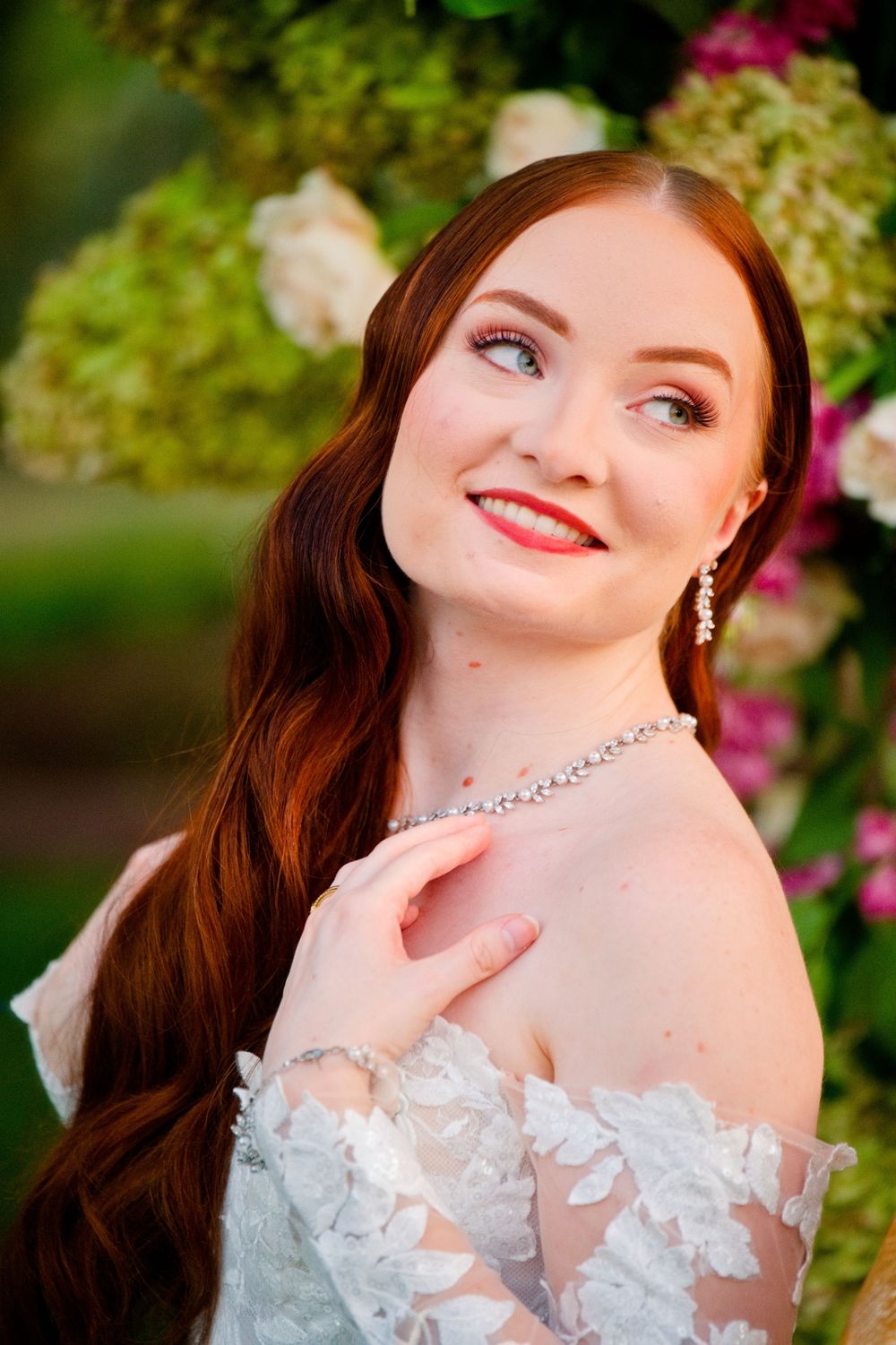 A bride in an elegant off-shoulder lace wedding gown poses against a backdrop of pink flowers and greenery outdoors.