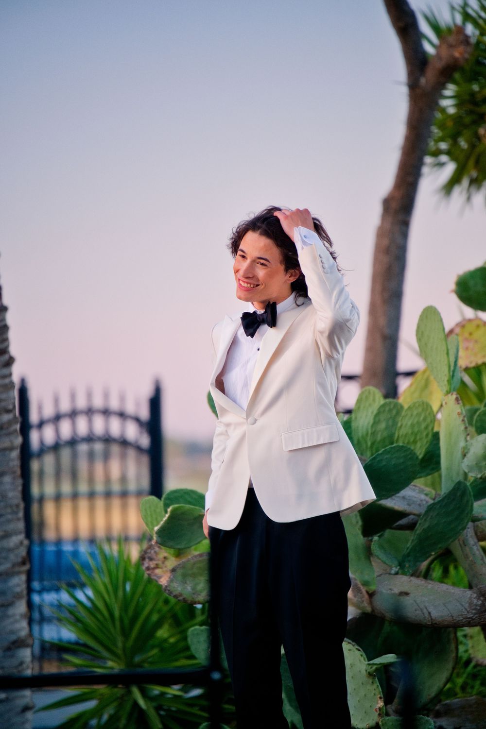 Someone in a classic white dinner jacket and black bow tie poses outdoors at sunset among tropical plants and palm trees.
