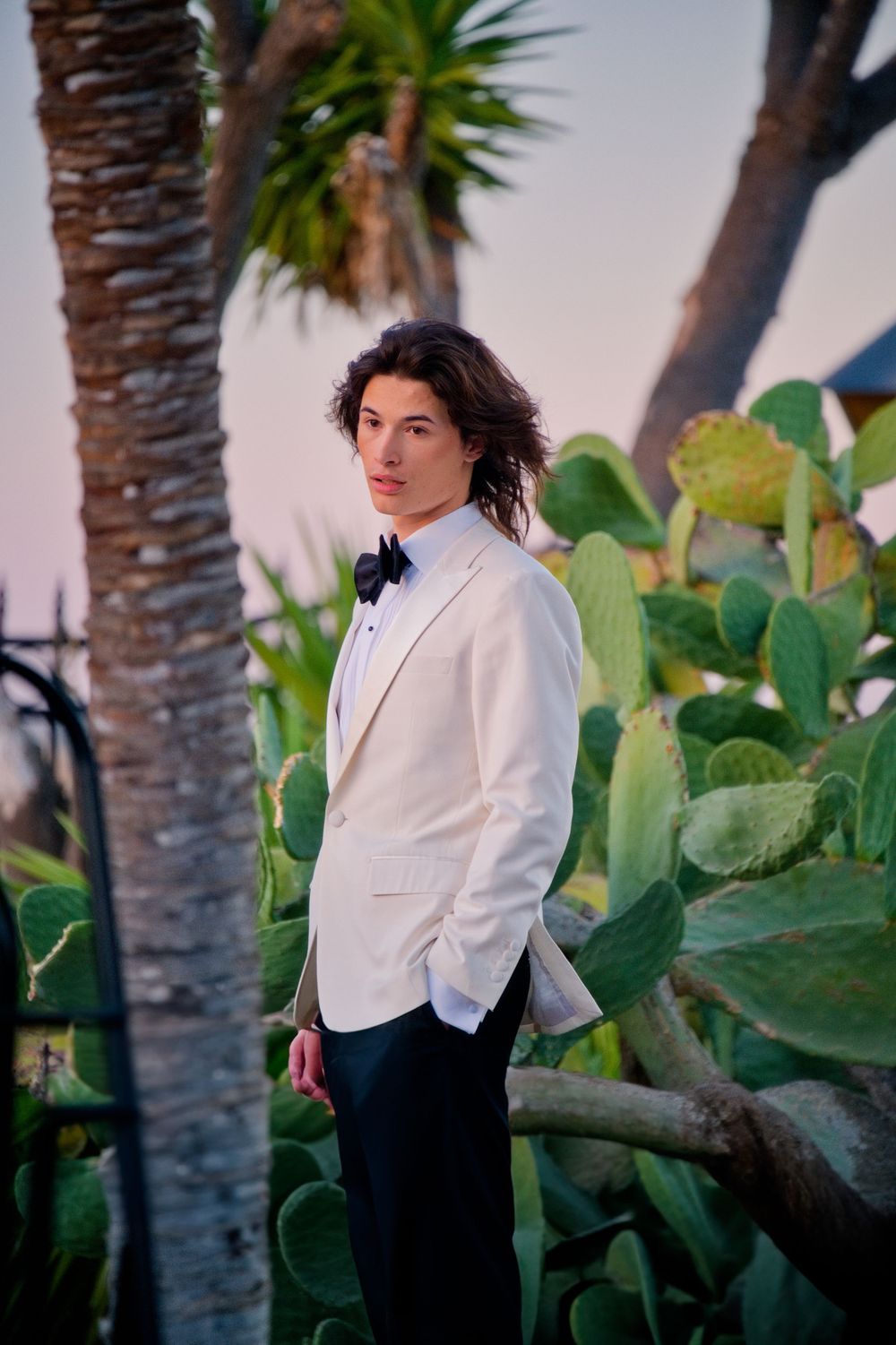 A formal portrait in a white blazer and bowtie against a tropical background with palm trees and cactus plants at sunset.