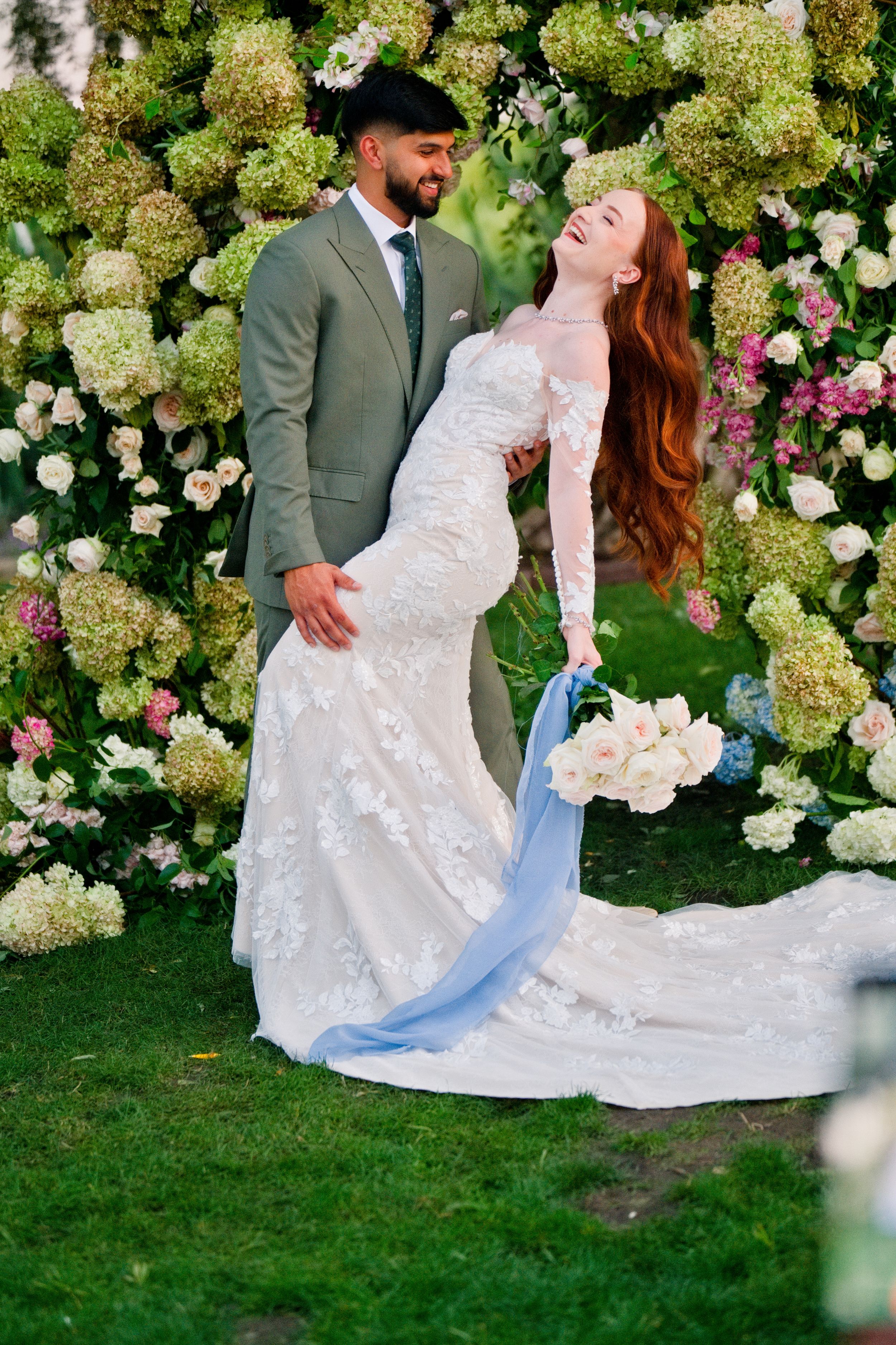 A joyful wedding scene with a couple posing among lush hydrangea bushes and roses in a garden setting.