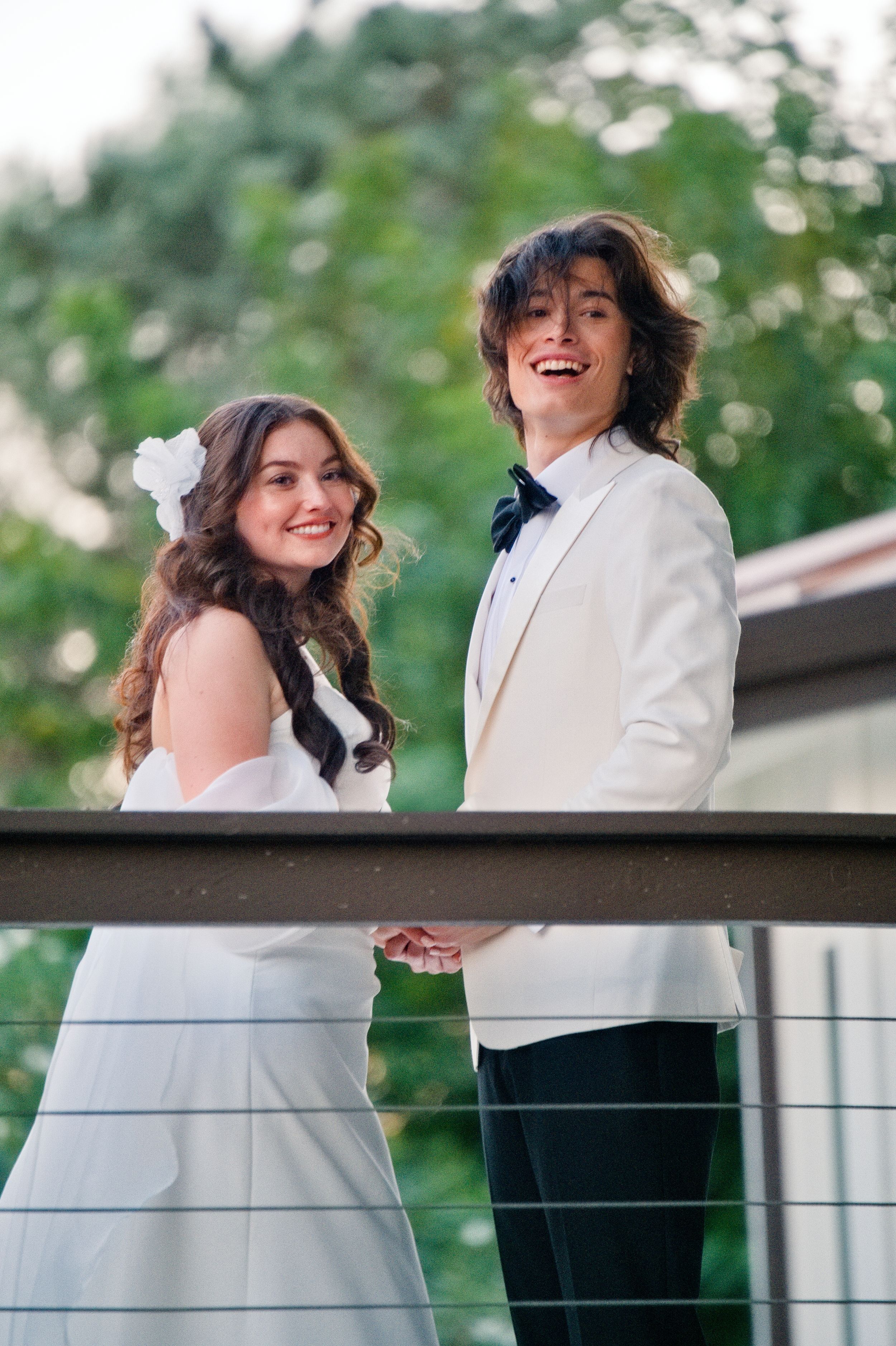 A happy couple in wedding attire standing outdoors on a deck with lush greenery in the background.