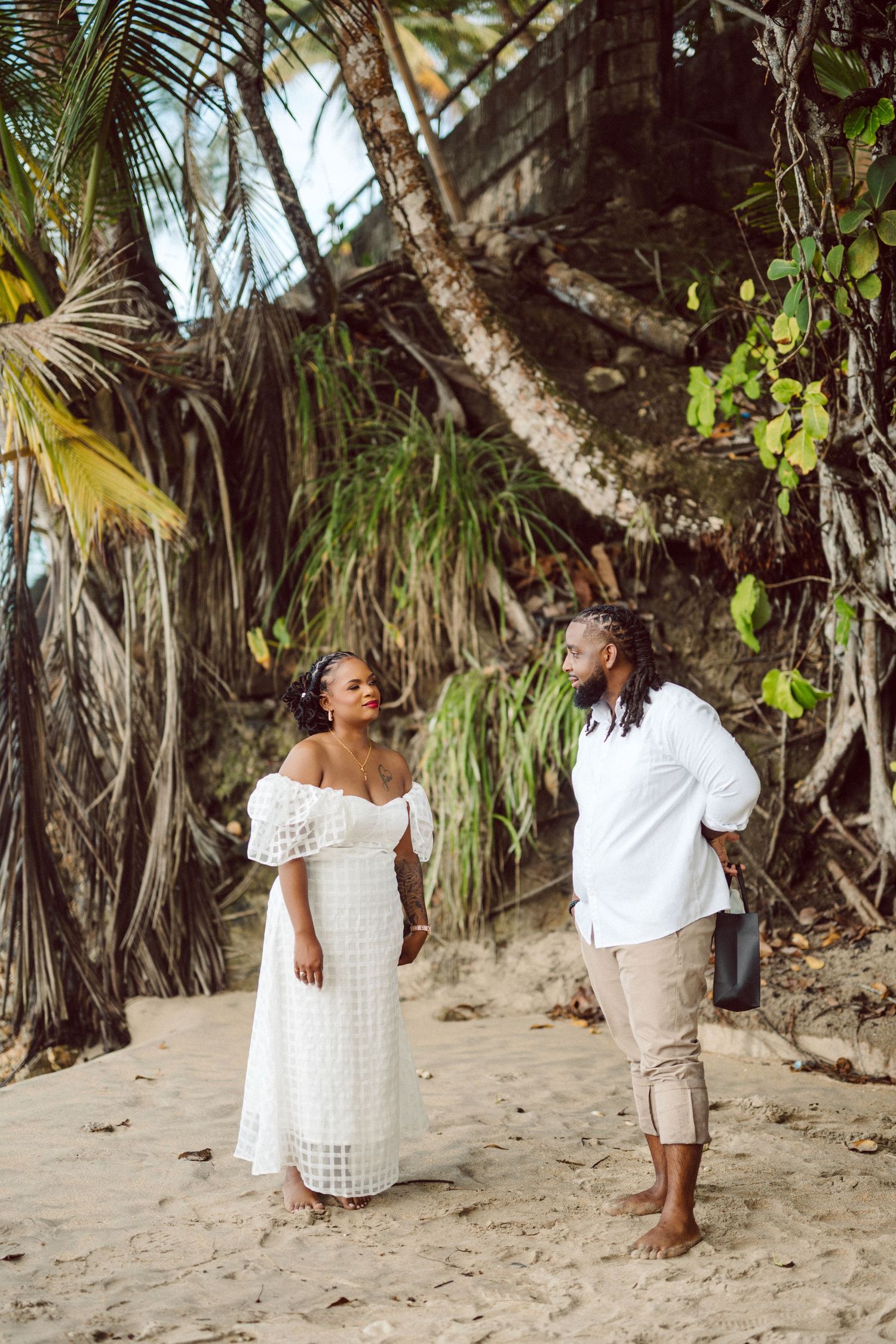 Two people standing on a tropical beach with palm trees and greenery in the background.