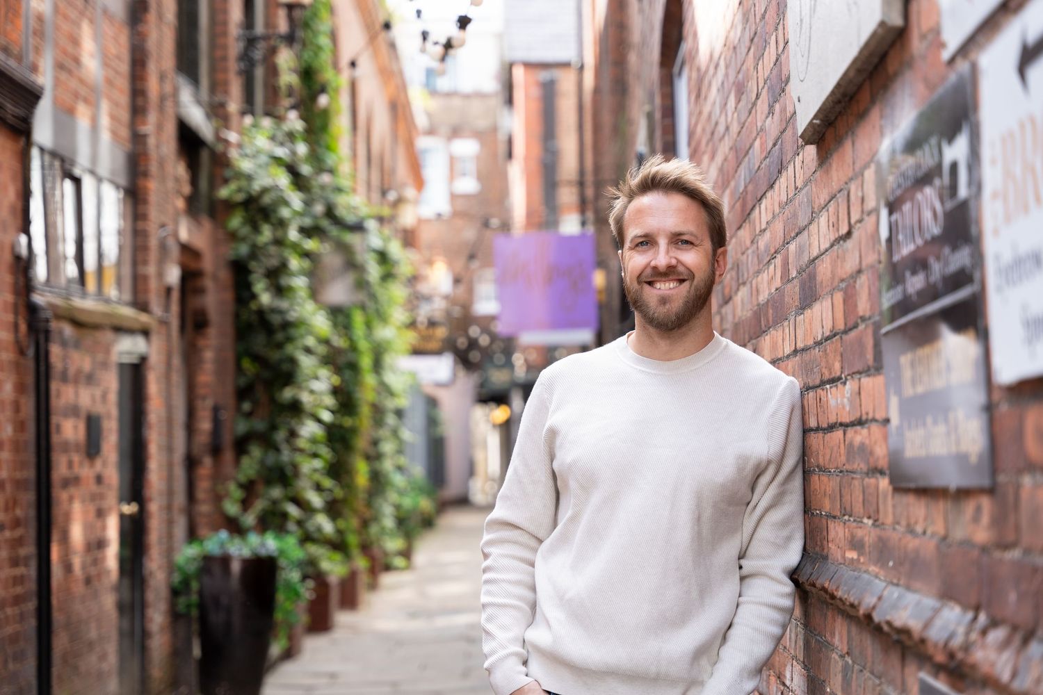 Life Coach and local businessman in cream sweater stands smiling in front of brick alley with ivy and architectural details.