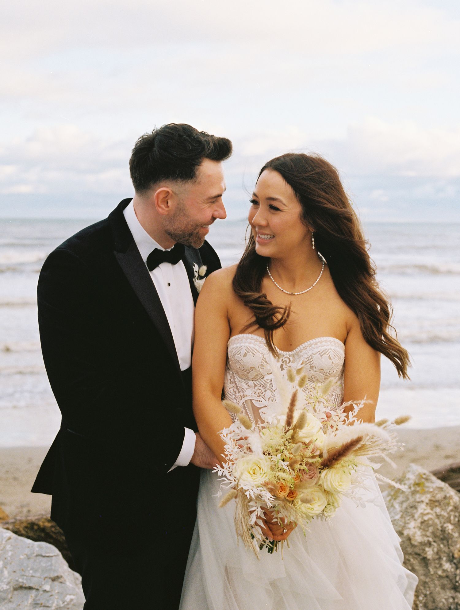 A newly married couple shares a romantic moment on a beach, with the bride holding a bouquet of cream-colored flowers.