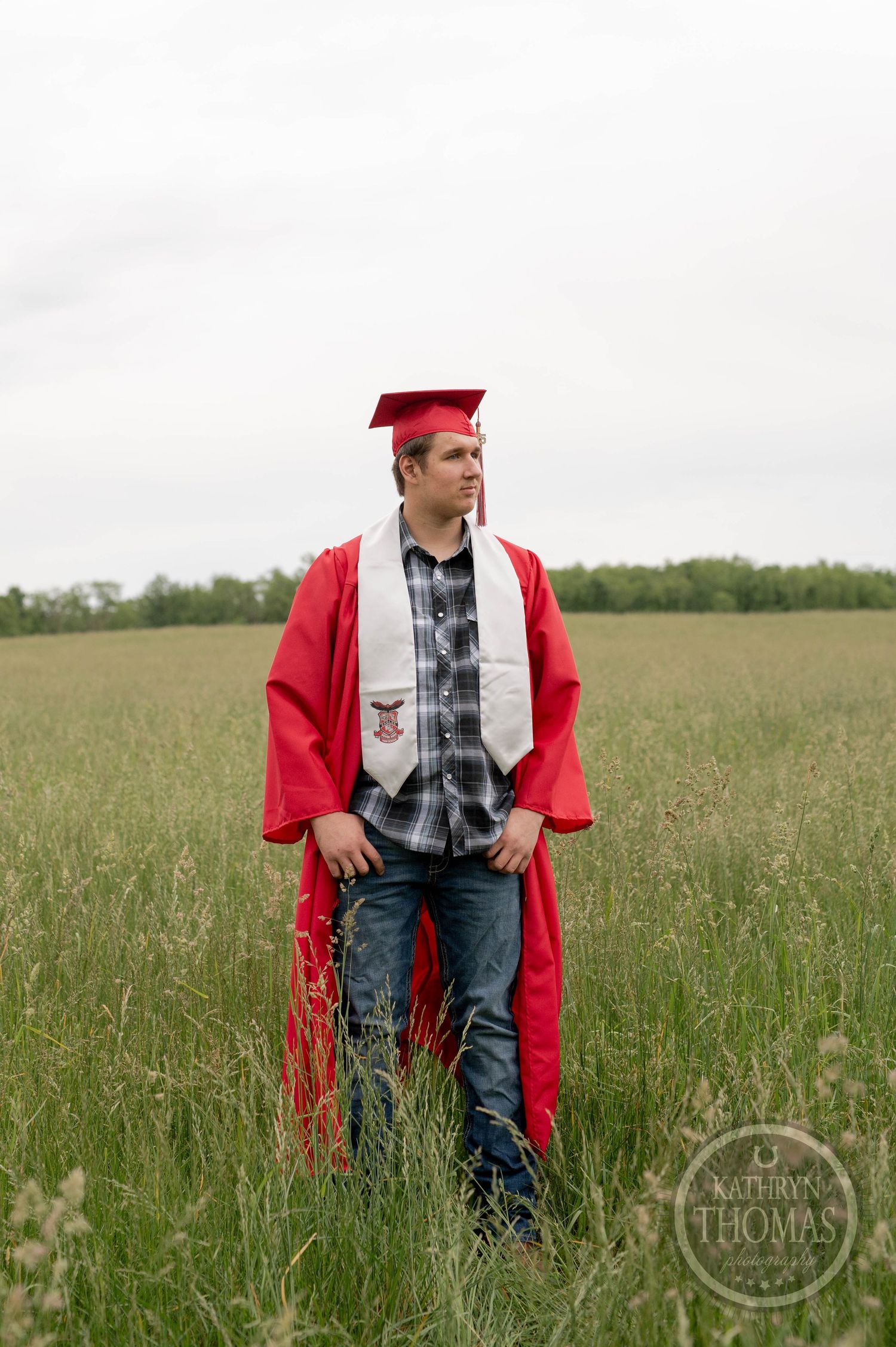 Graduate in red cap and gown stands in tall grass field wearing plaid shirt and jeans during outdoor photo session.