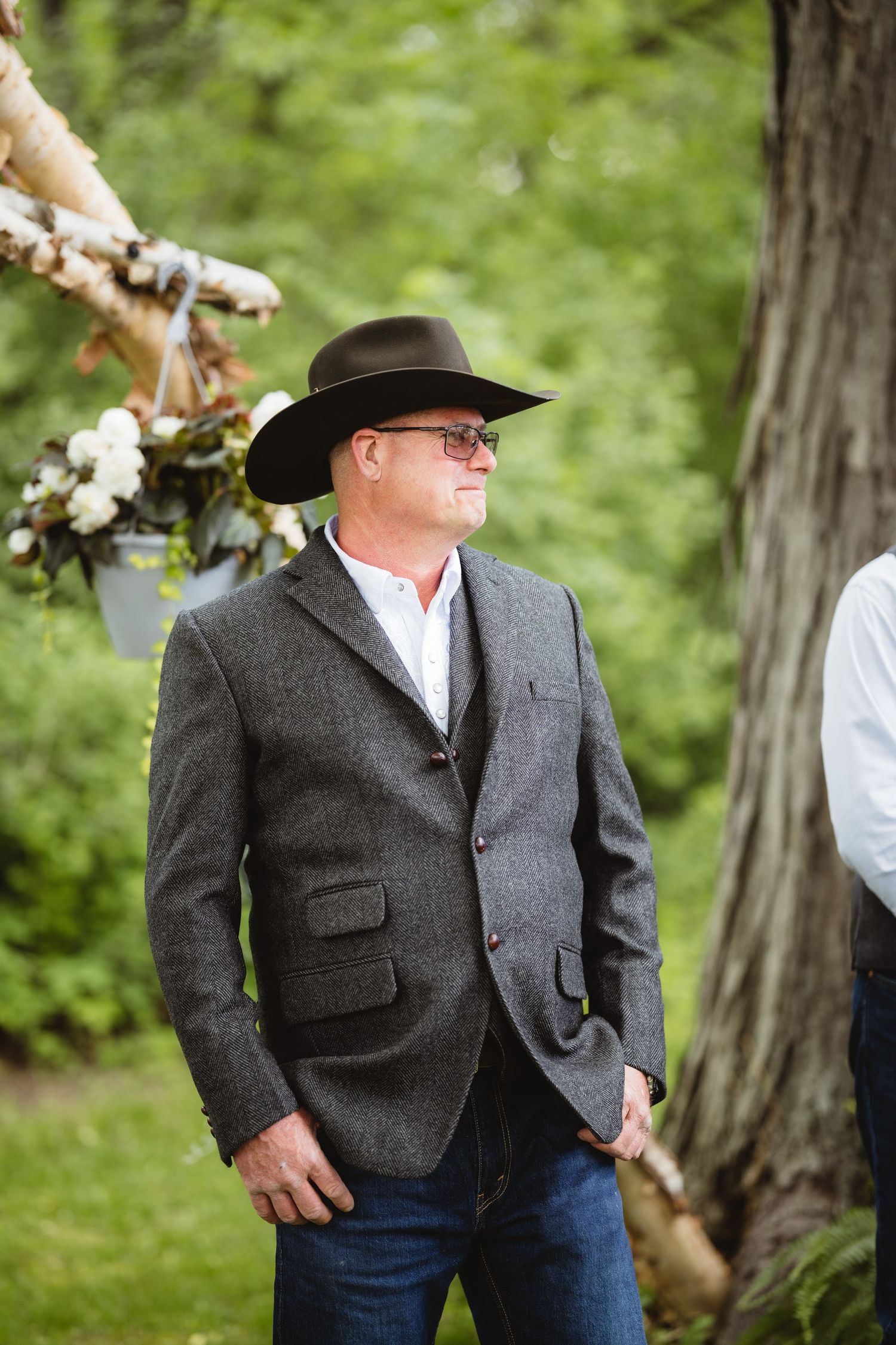 Person wearing a gray suit jacket and cowboy hat stands in front of green foliage outdoors.