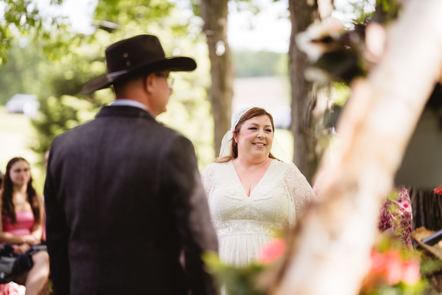 A couple during their outdoor wedding ceremony with blurred figures in the background on a sunny day.