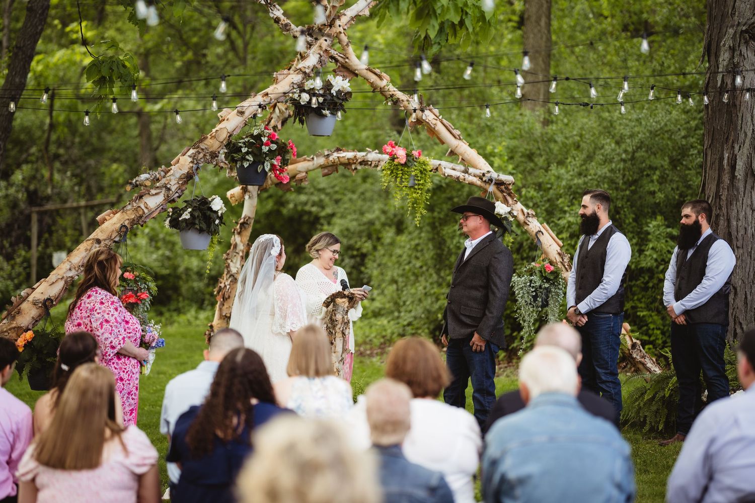 Rustic outdoor wedding ceremony under a decorated birch arch with guests seated in a woodland setting.
