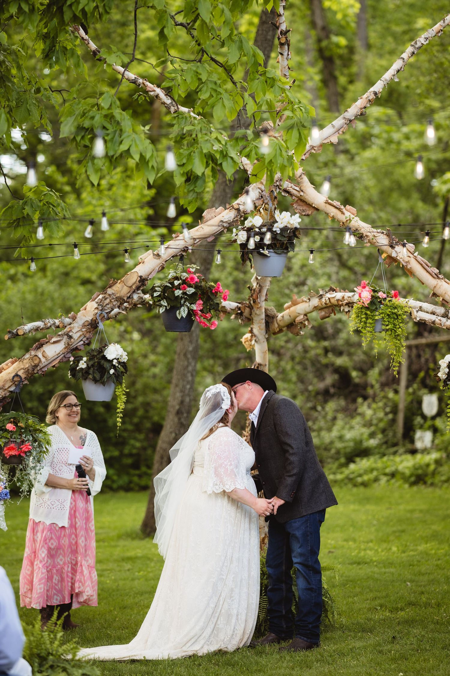 A rustic outdoor wedding ceremony takes place under a decorated branch arch with hanging floral arrangements in a garden setting.