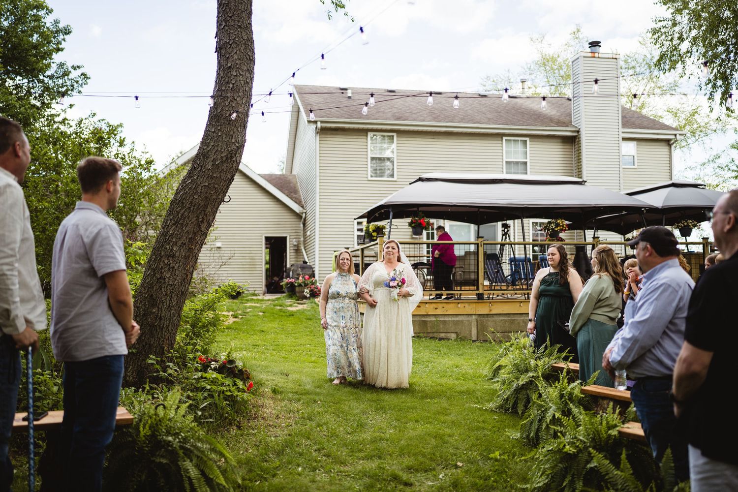 An outdoor backyard wedding ceremony takes place in front of a white two-story house with guests gathered on the lawn.