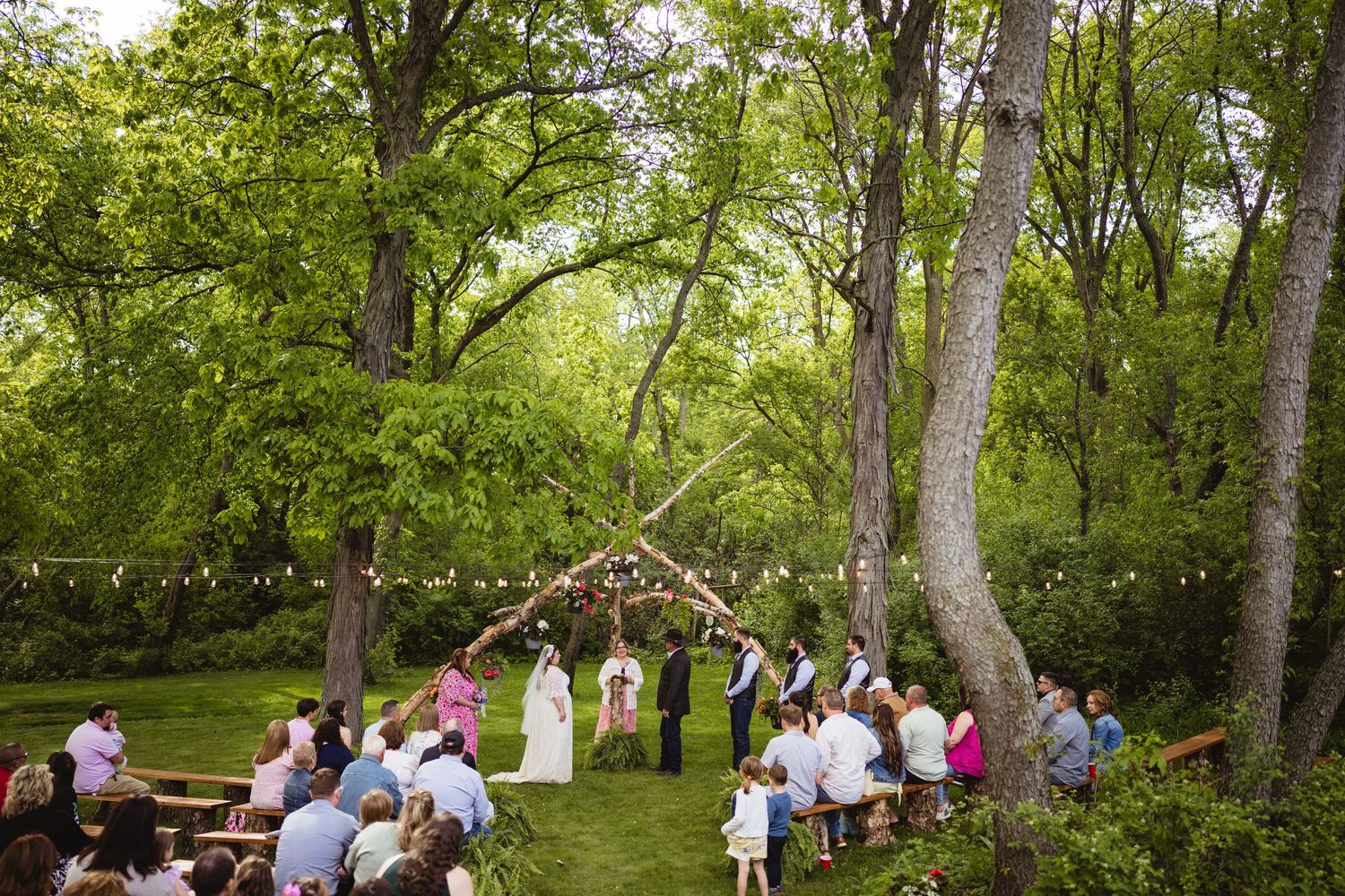 A rustic outdoor wedding ceremony in a lush green forest with guests seated beneath string lights and tall trees.