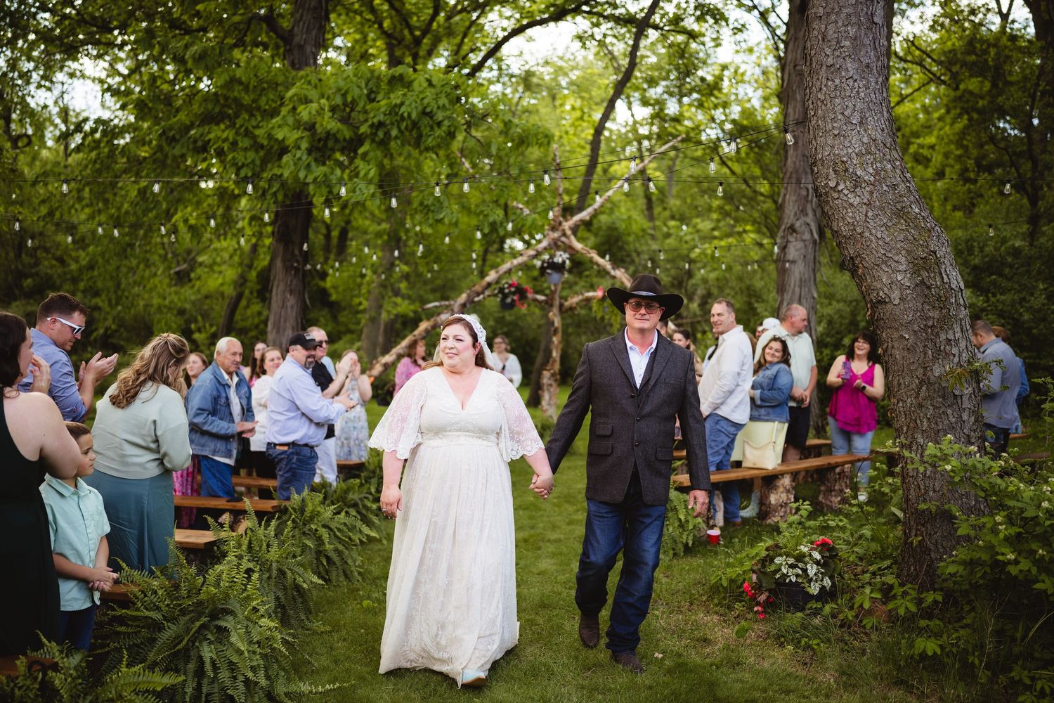 A rustic outdoor wedding ceremony takes place under tall trees with guests seated on wooden benches in a garden setting.