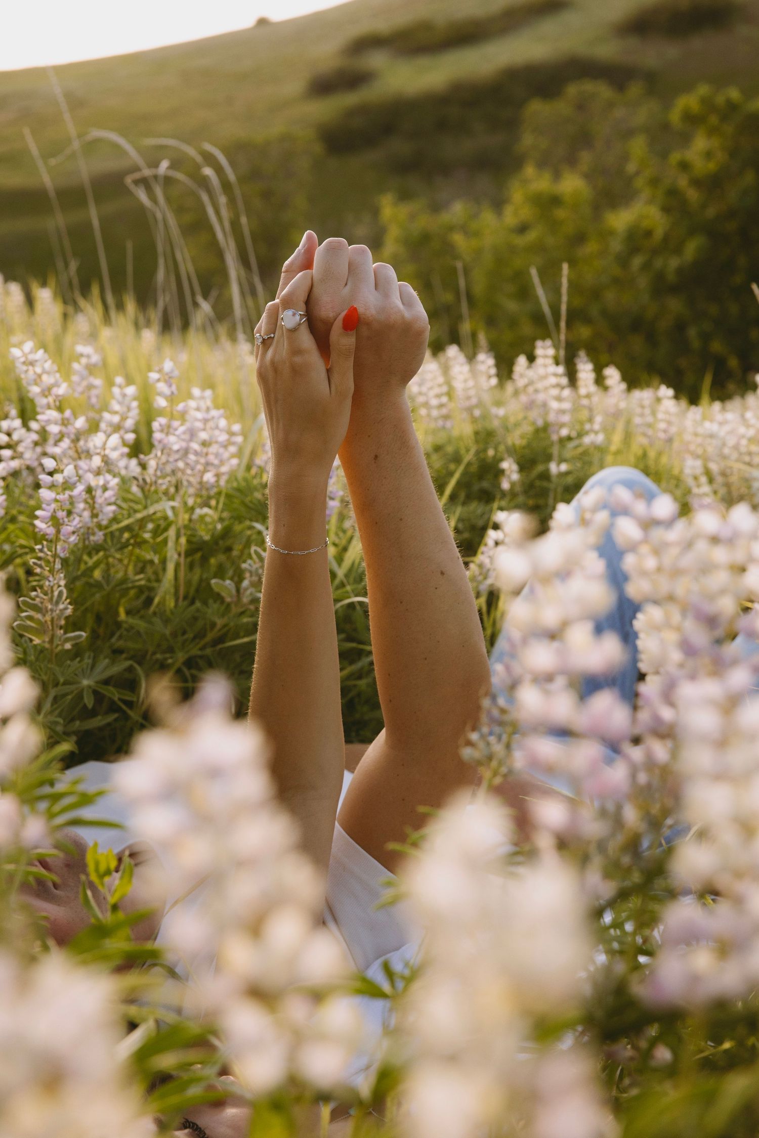 Raised hands reach skyward among delicate purple lupine flowers in a sunlit meadow during golden hour.