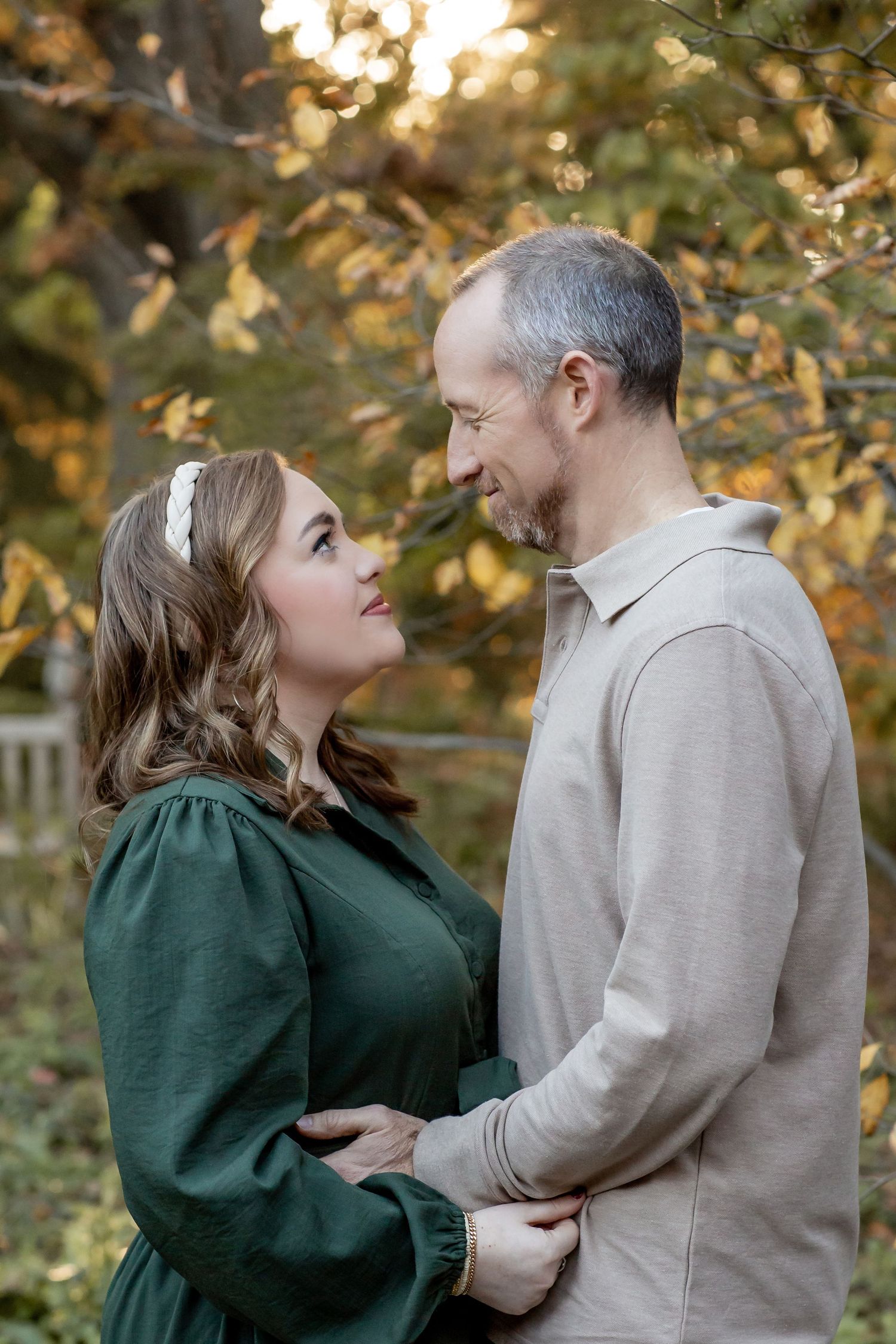 Couple gazing at each other tenderly outdoors, woman in green dress and man in beige sweater among fall foliage.
