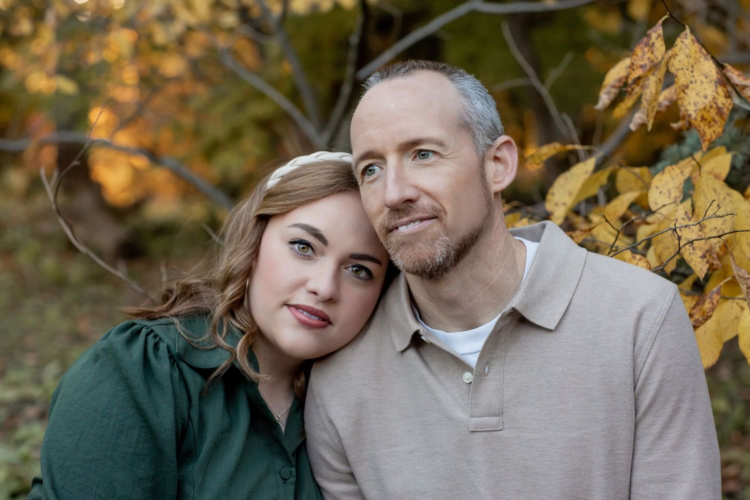 Couple posing closely together outdoors surrounded by golden autumn leaves, woman leaning on mans shoulder.
